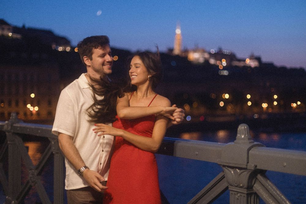 Smiling couple dancing on a bridge at twilight with illuminated Budapest skyline.
