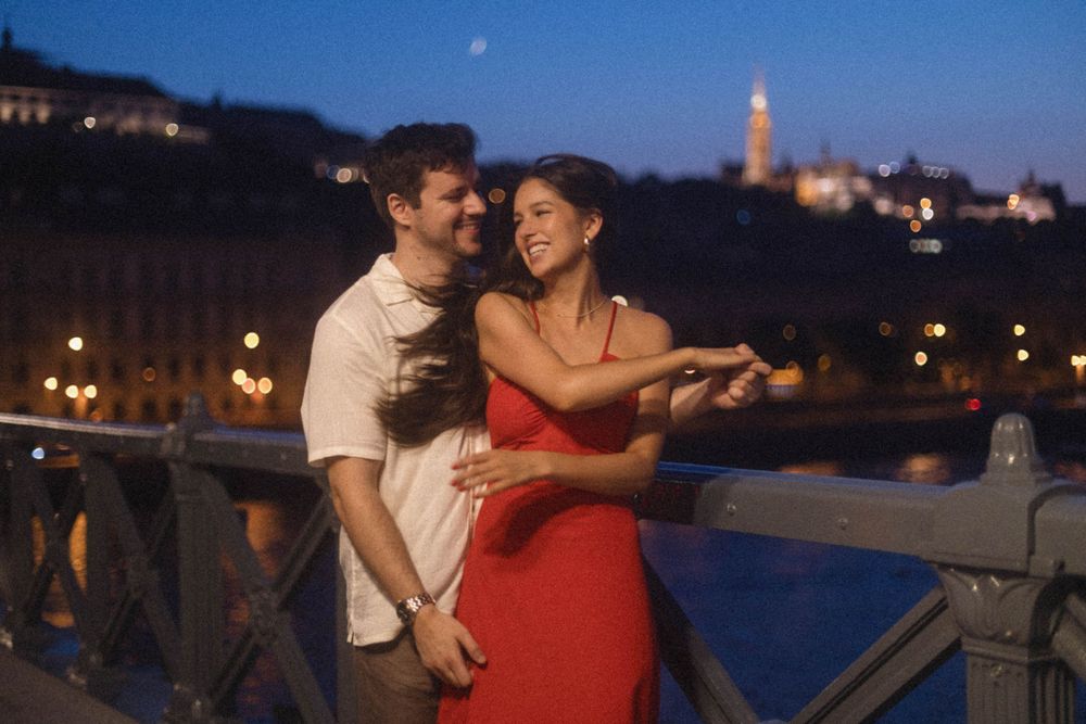 Happy couple holding hands on a bridge at dusk with Budapest landmarks in background.
