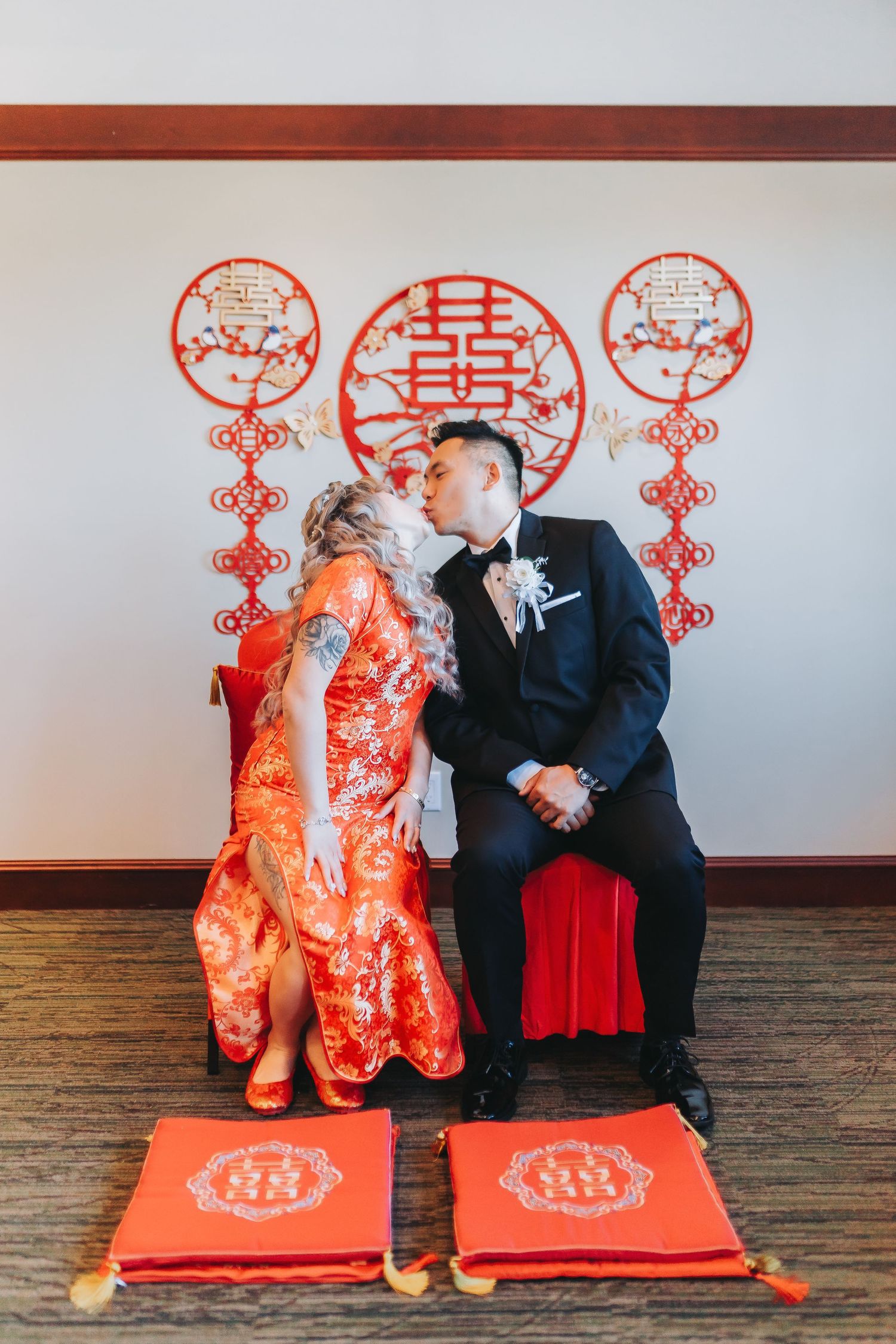 Wedding couple sitting on red cushions with traditional Chinese double happiness symbols on wall behind them.