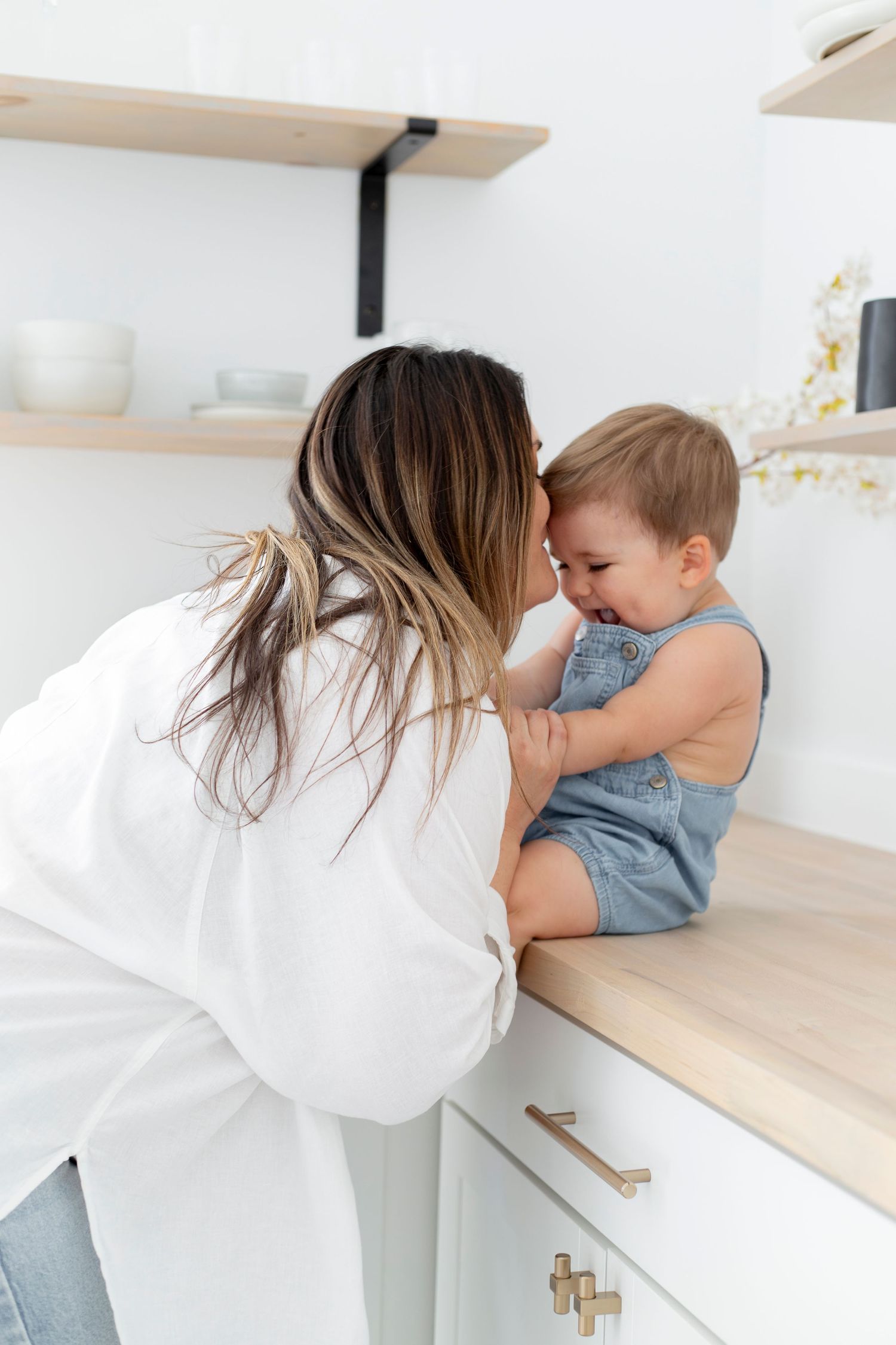 A tender moment between parent and laughing toddler sitting on a kitchen counter in a bright white modern kitchen.