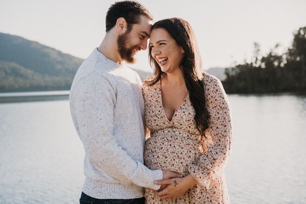 pregnant couple standing leaning into each other and laughin with hands meeting on pregnant belly at Lake Kaniere on the West Coast