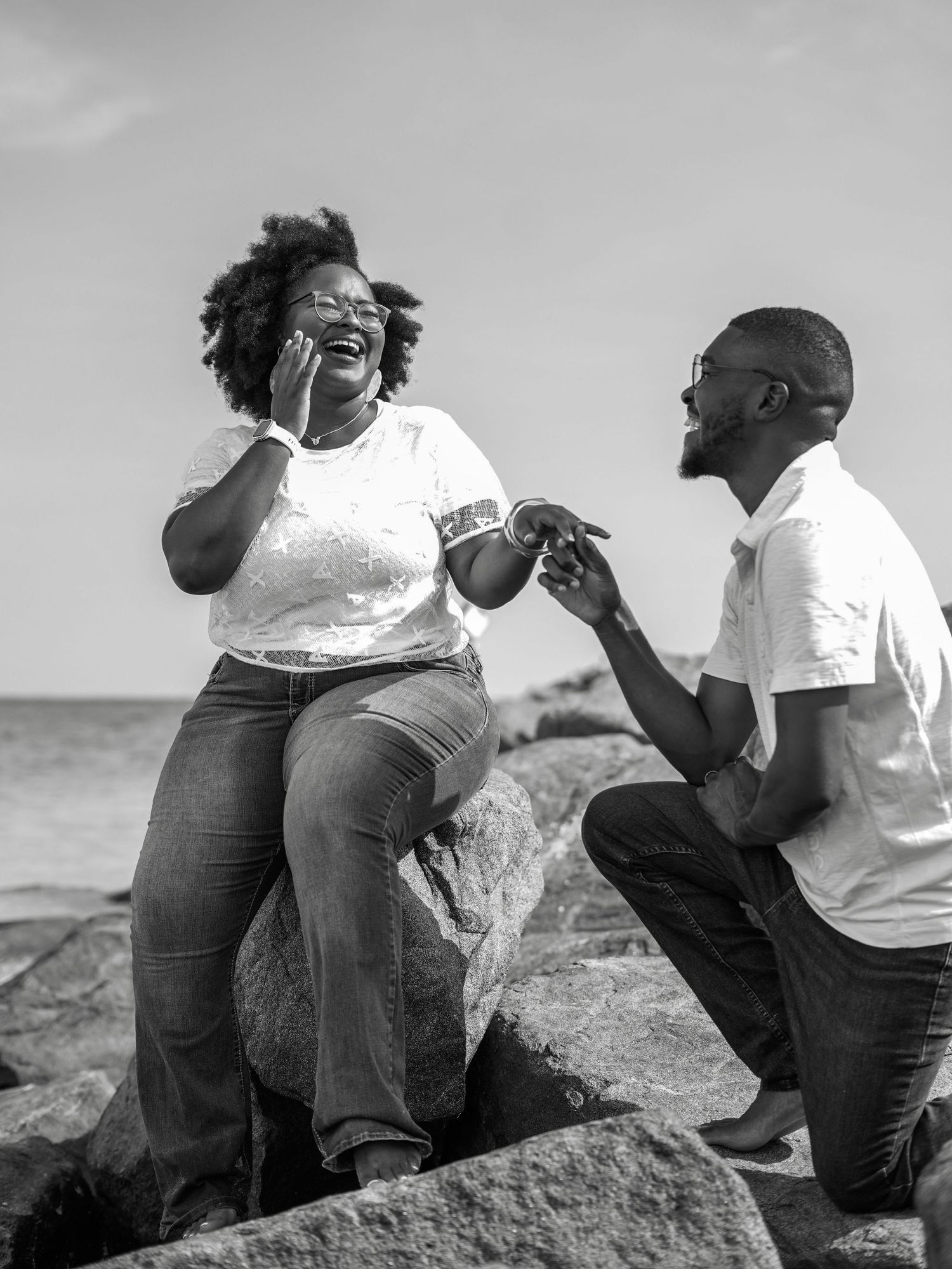 A surprise proposal moment captured on coastal rocks in black and white photography.
