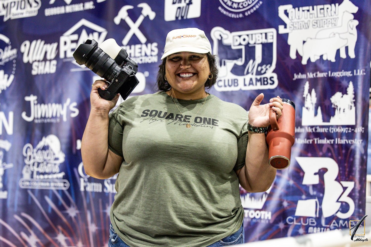 A smiling woman holds a camera and a Stanley cup in front of a Square One-branded banner at an event.