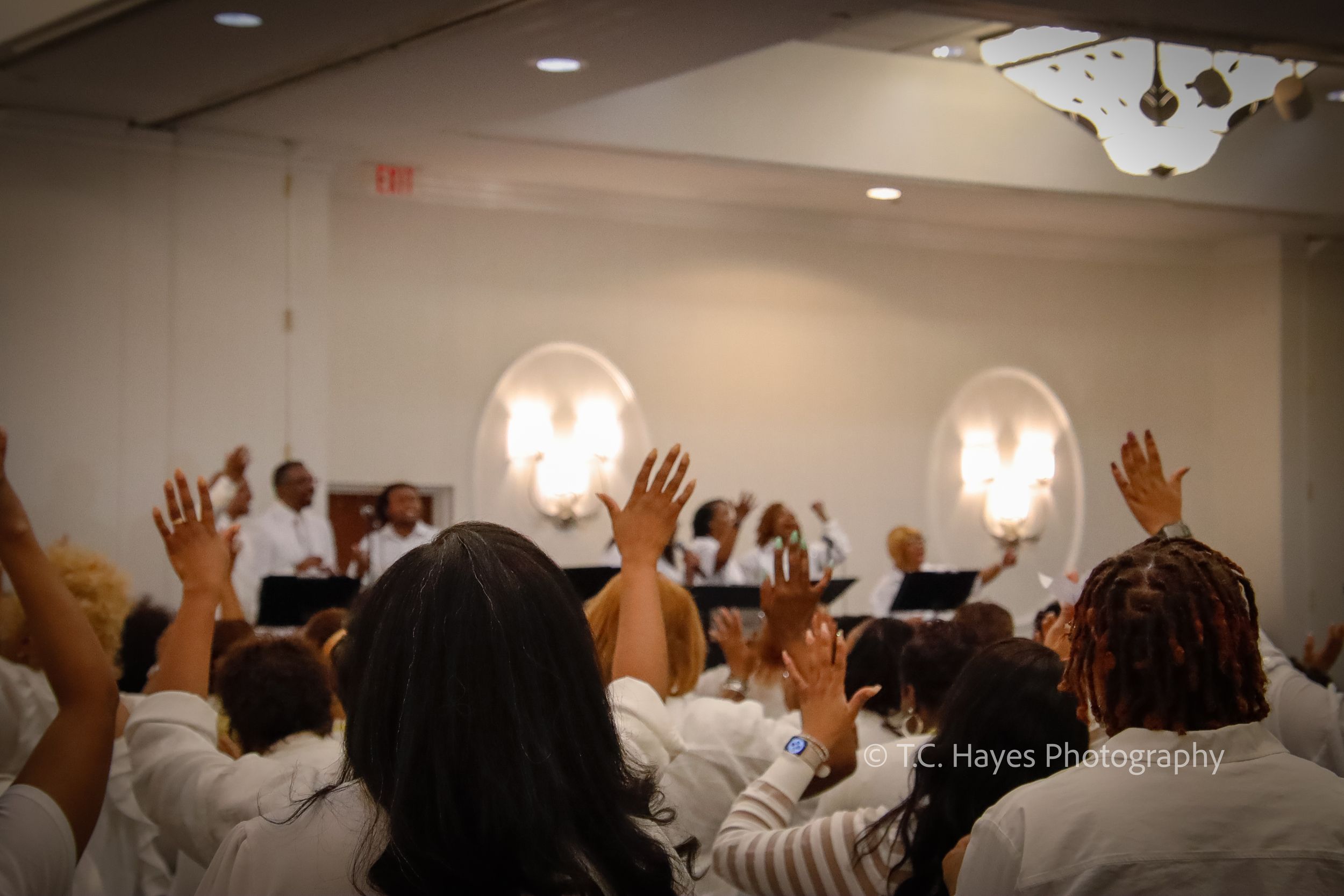 Crowd of people in white raising hands during a worship service with illuminated wall sconces.