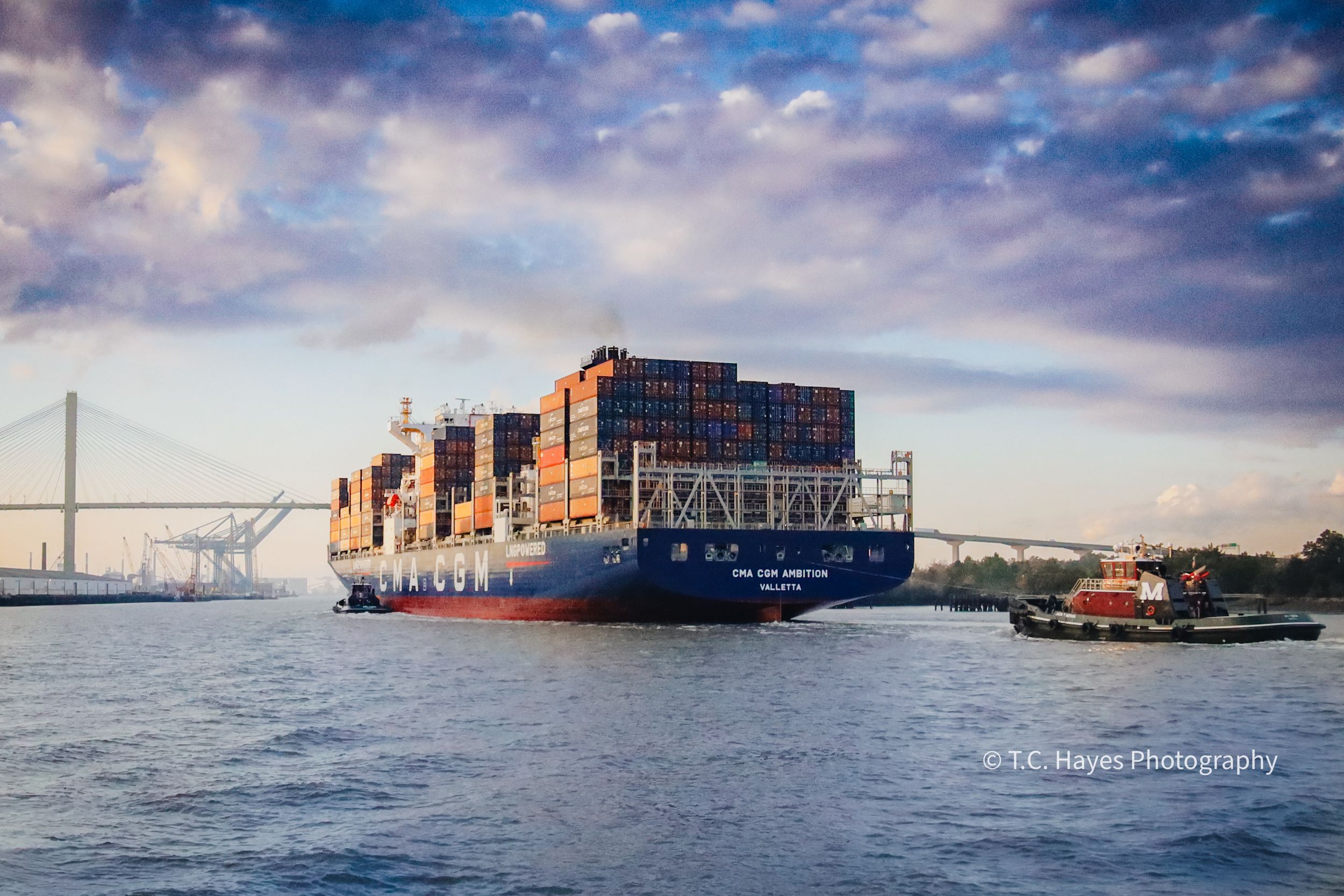 Large cargo container ship passing under bridge in harbor during golden hour.