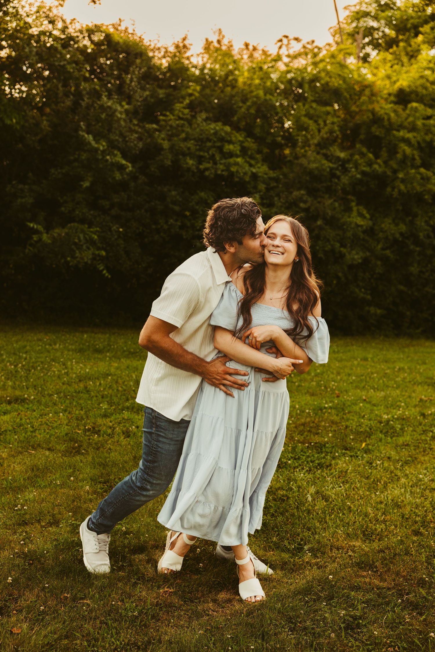 A couple embraces on a lush green lawn at sunset, sharing a tender moment against a backdrop of golden-lit foliage.