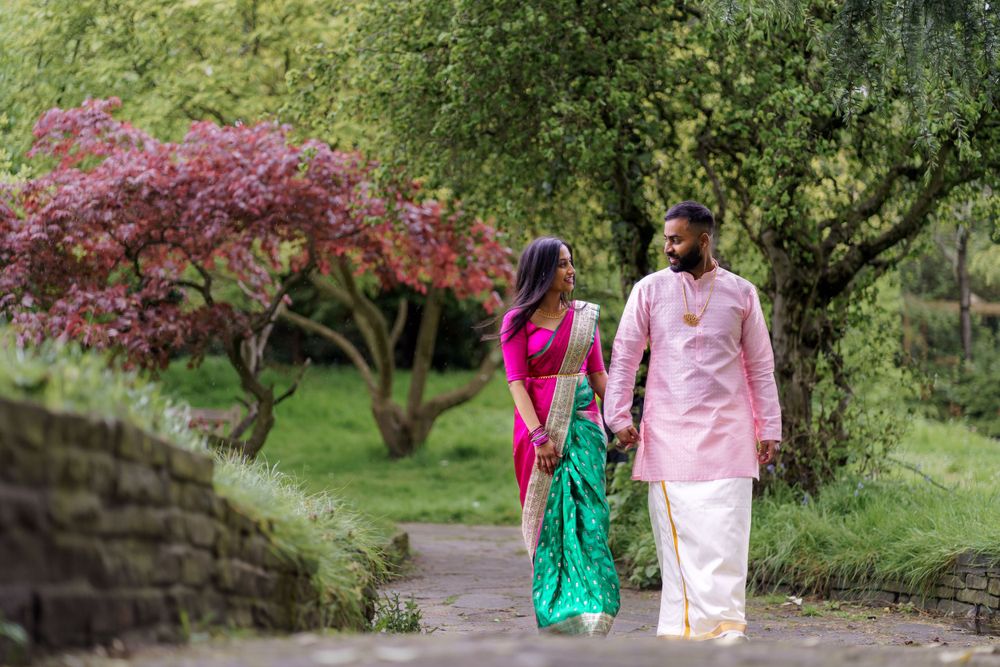 Two people in traditional Indian attire walk along a garden path beneath pink flowering trees.