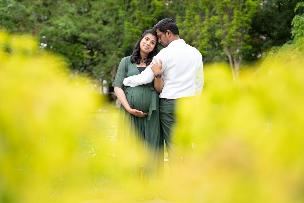 A couple in green and white outfits embrace lovingly in a garden setting, framed by soft yellow blooms in the foreground.