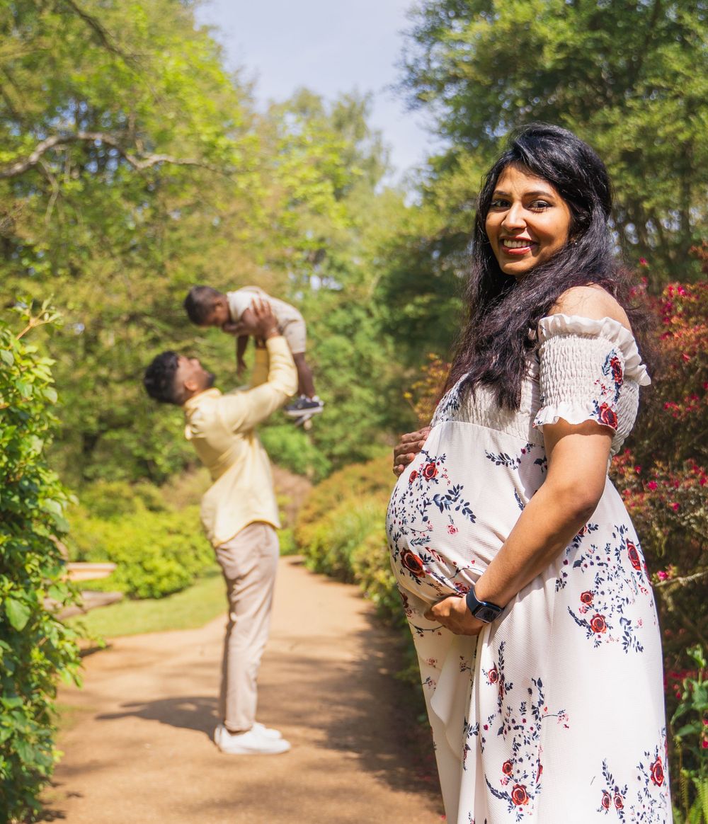 A pregnant woman in a floral dress stands on a garden path while someone lifts a child in the background.