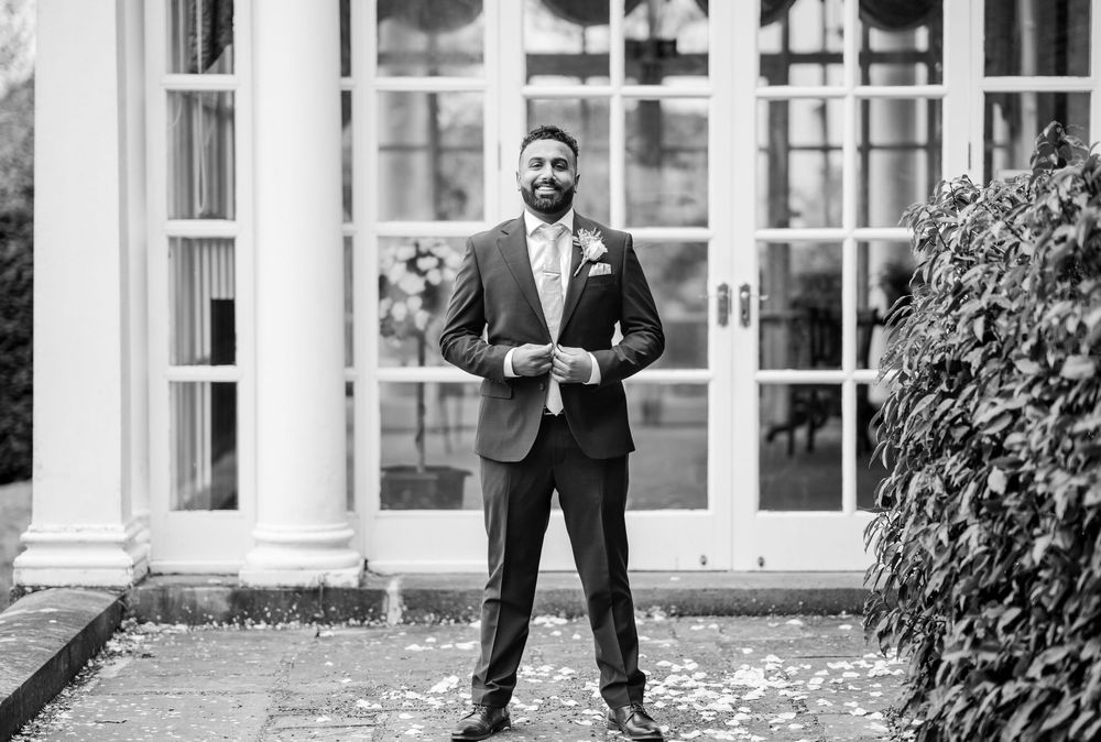 A person in a dark suit stands smiling outdoors with hands in pockets in front of an elegant building entrance.