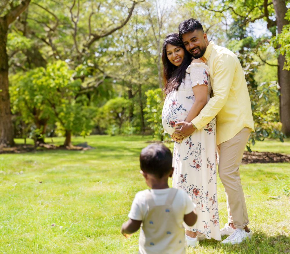 A couple in a floral dress and yellow shirt embrace in a sunny park while a child plays in the foreground.
