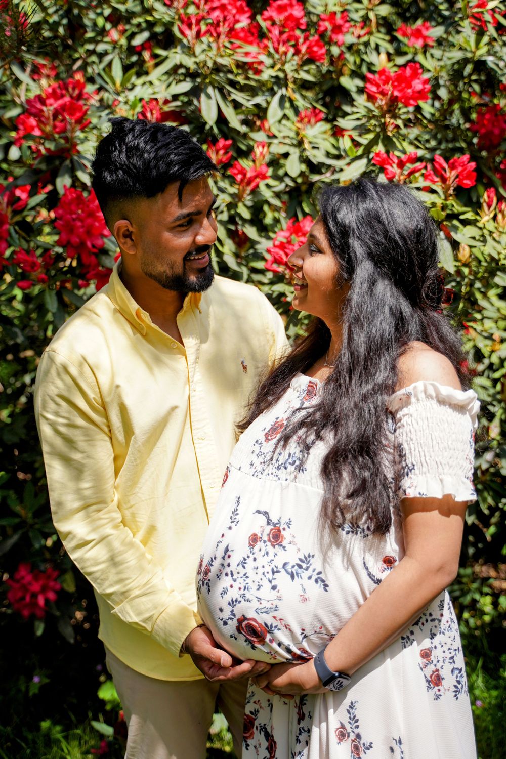 A couple share an intimate moment in front of vibrant red flowers and greenery on a sunny day as part of their maternity shoot.