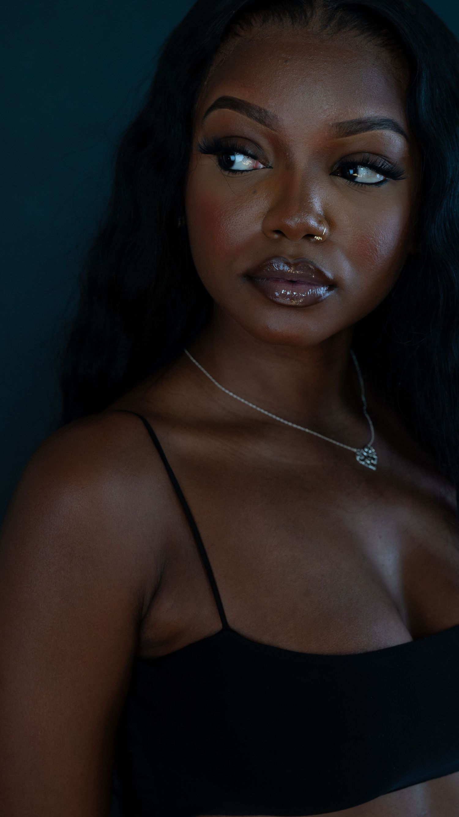 Fashion model poses in black top against dark background with delicate necklace and glamorous makeup.