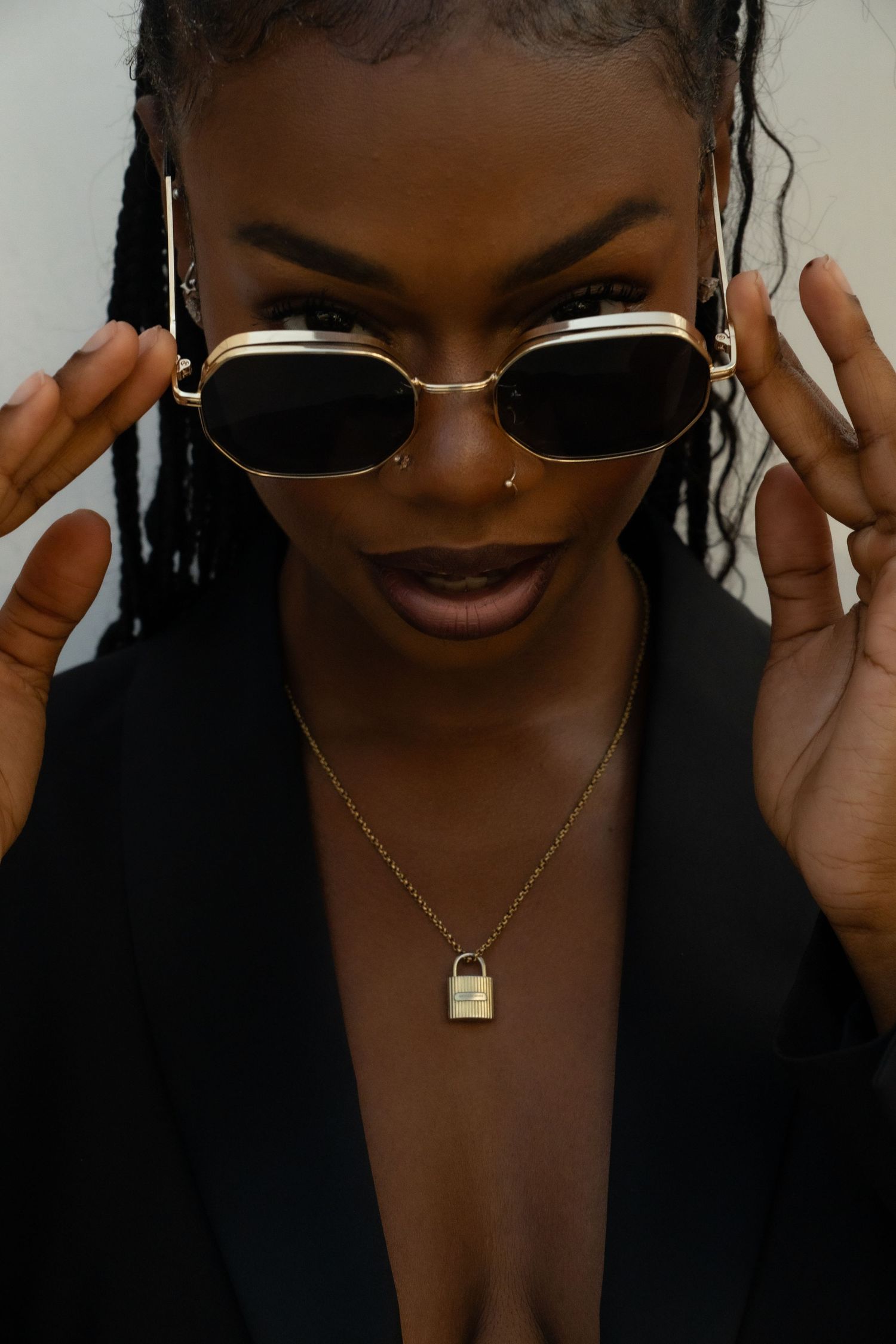 Stylish fashion portrait featuring retro sunglasses, gold lock necklace and black blazer in moody lighting.