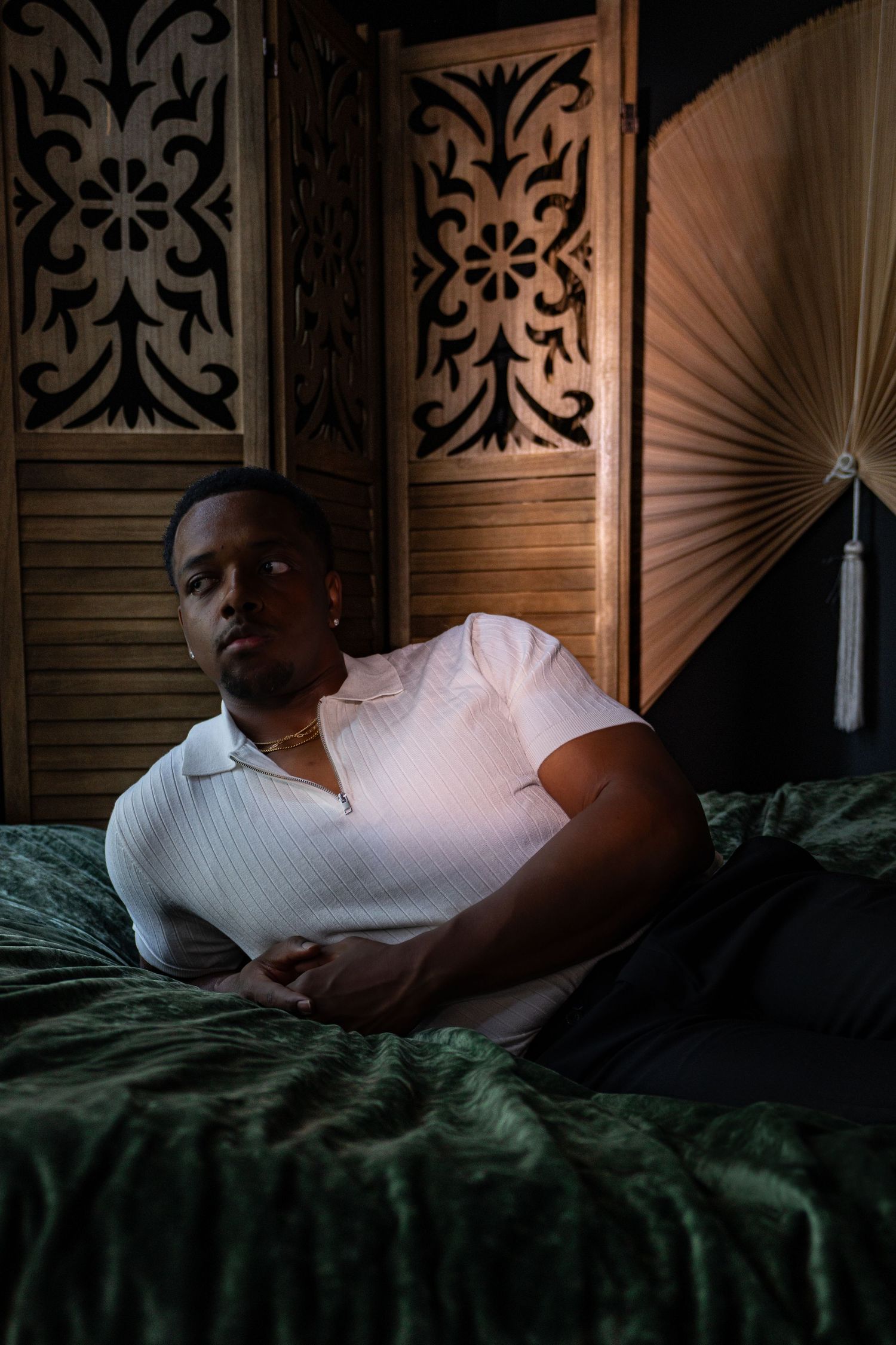 Young man in white polo shirt reclining on green velvet bed with ornate wooden screen backdrop.