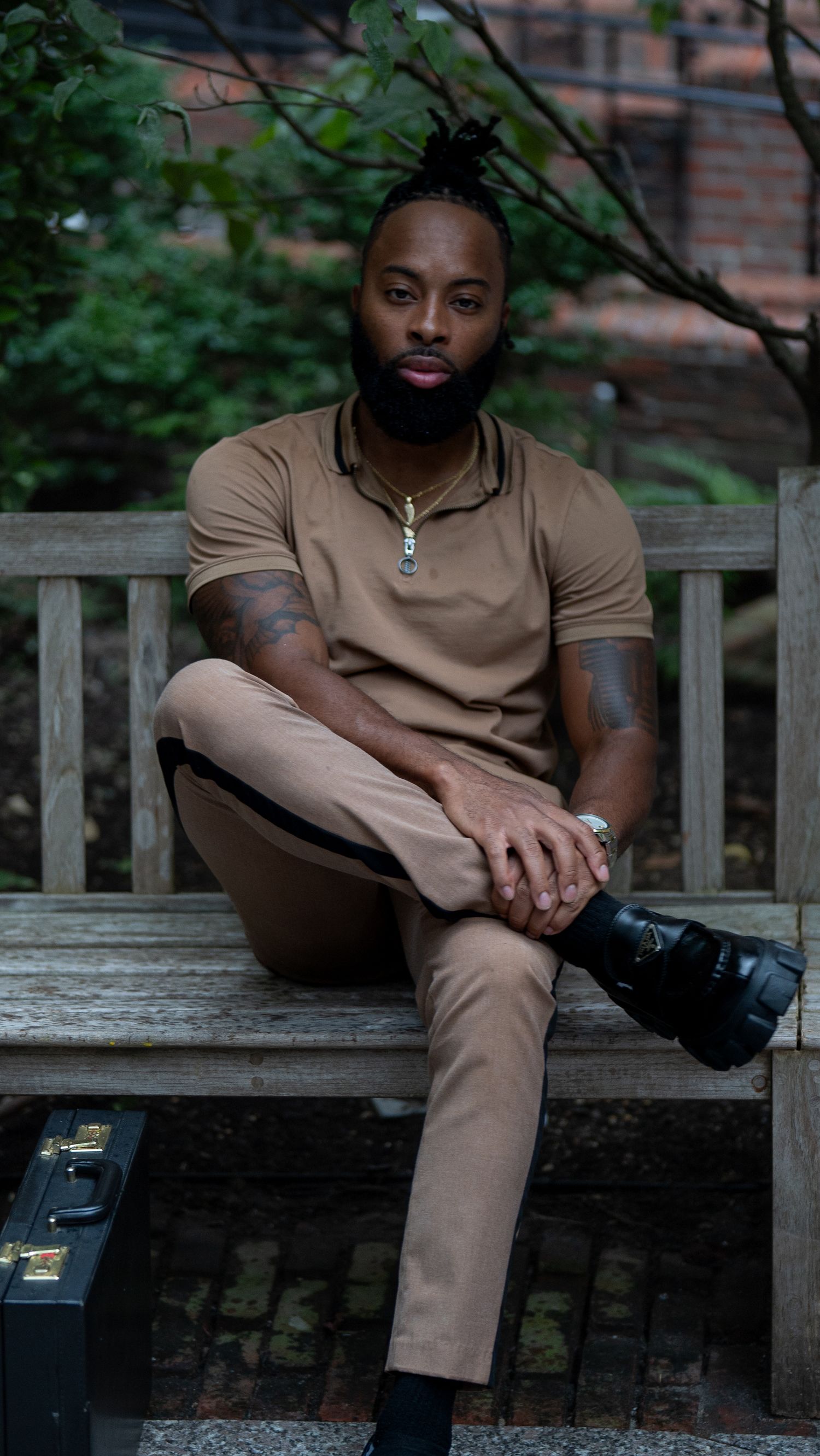 Fashionable person in brown outfit sitting on wooden bench with camera and briefcase.