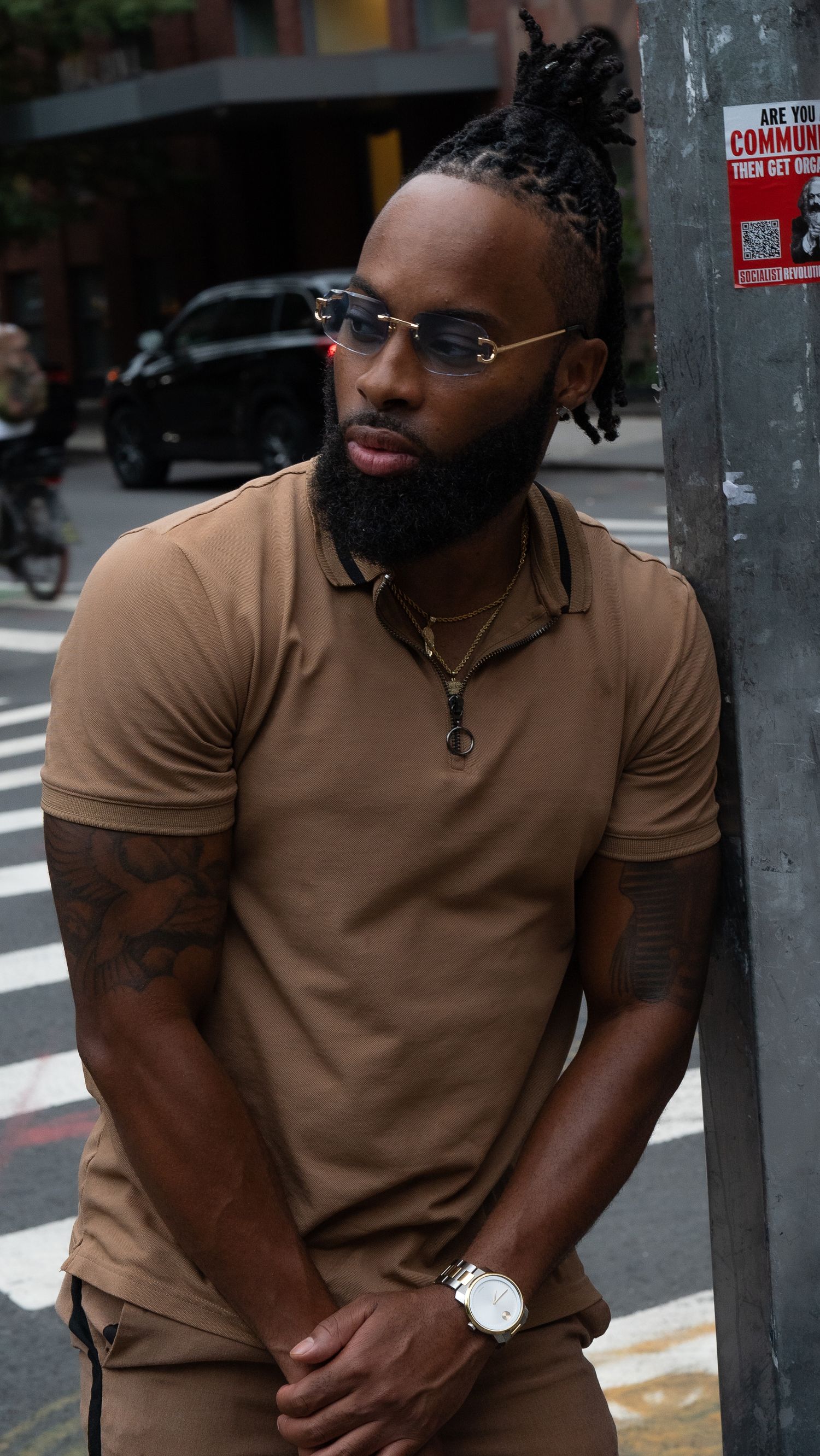 Stylish individual in brown polo with glasses standing at urban intersection.