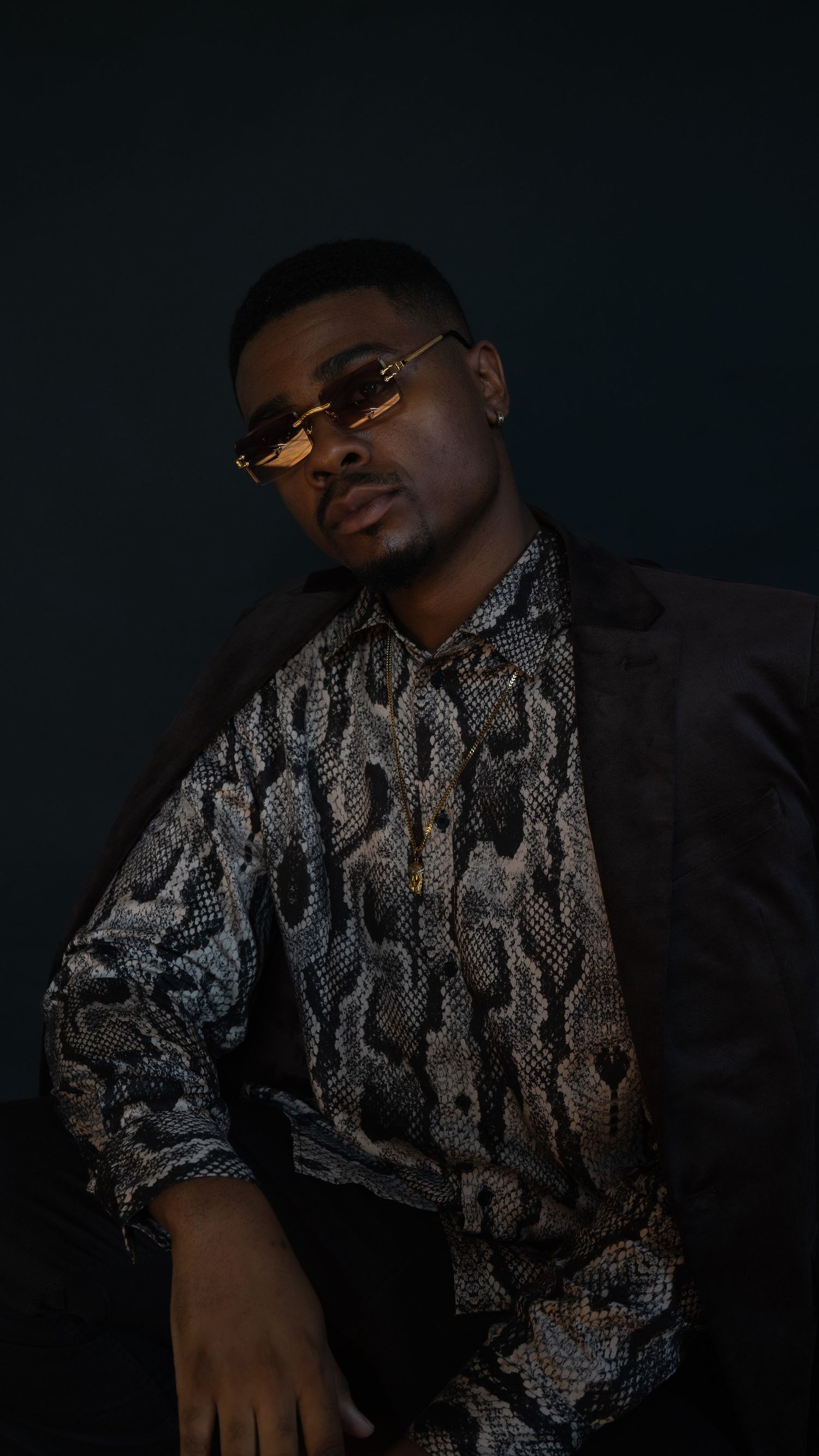 Stylish portrait in snakeskin print shirt and sunglasses against dark background.