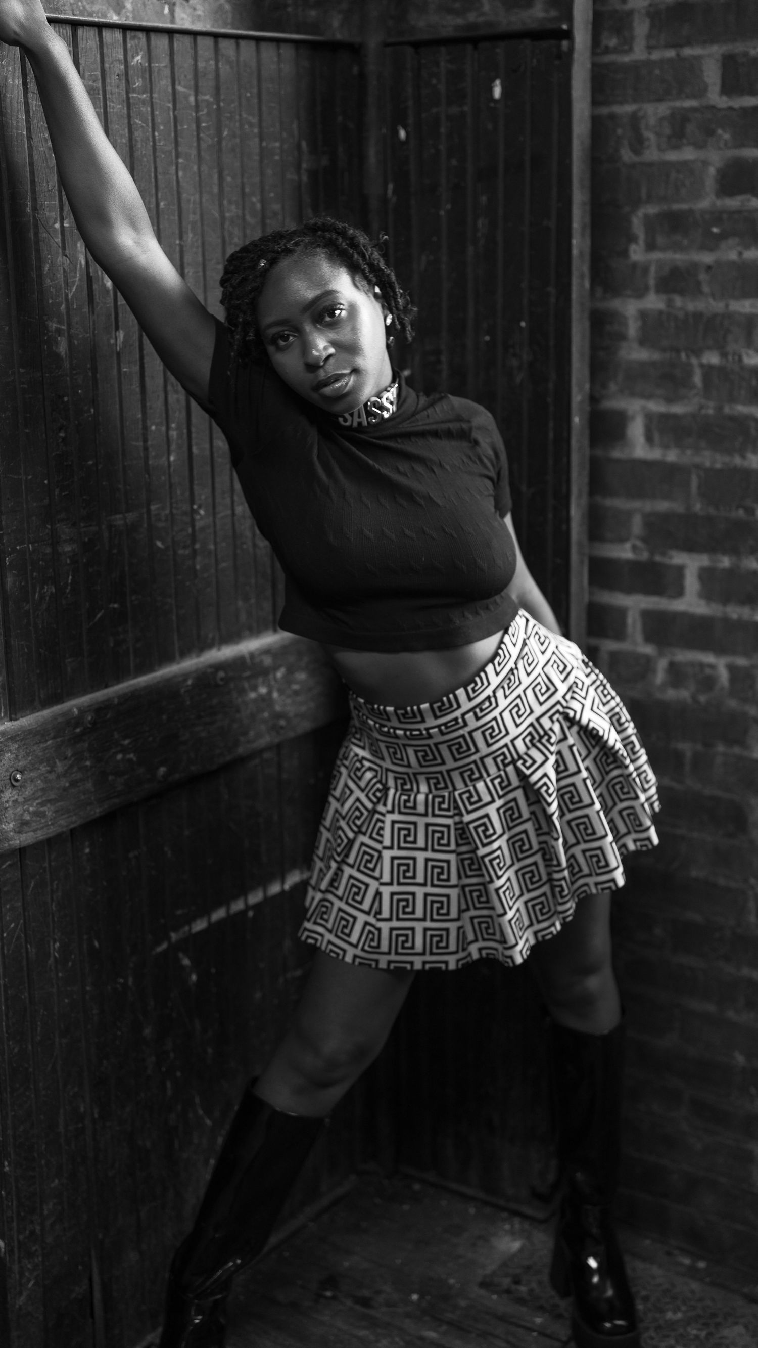 Dancer in black crop top and patterned skirt strikes dynamic pose against brick wall in black and white photo.