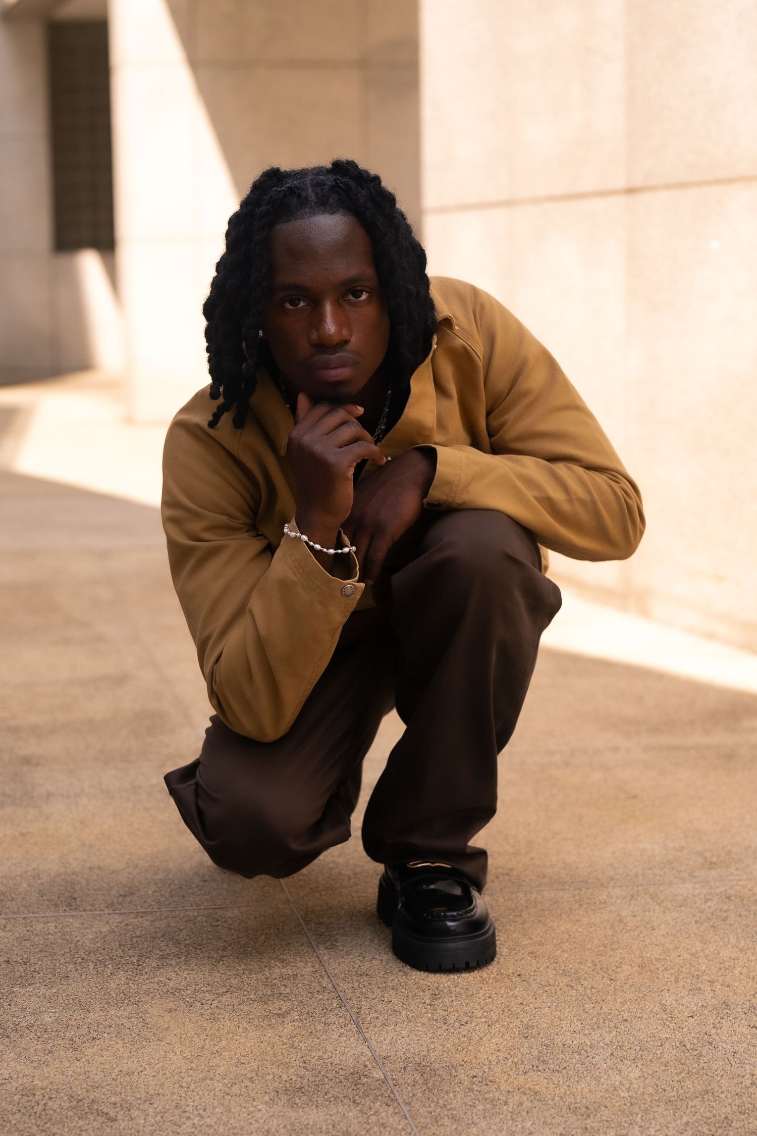Urban fashion portrait of person in brown jacket crouching in architectural setting.
