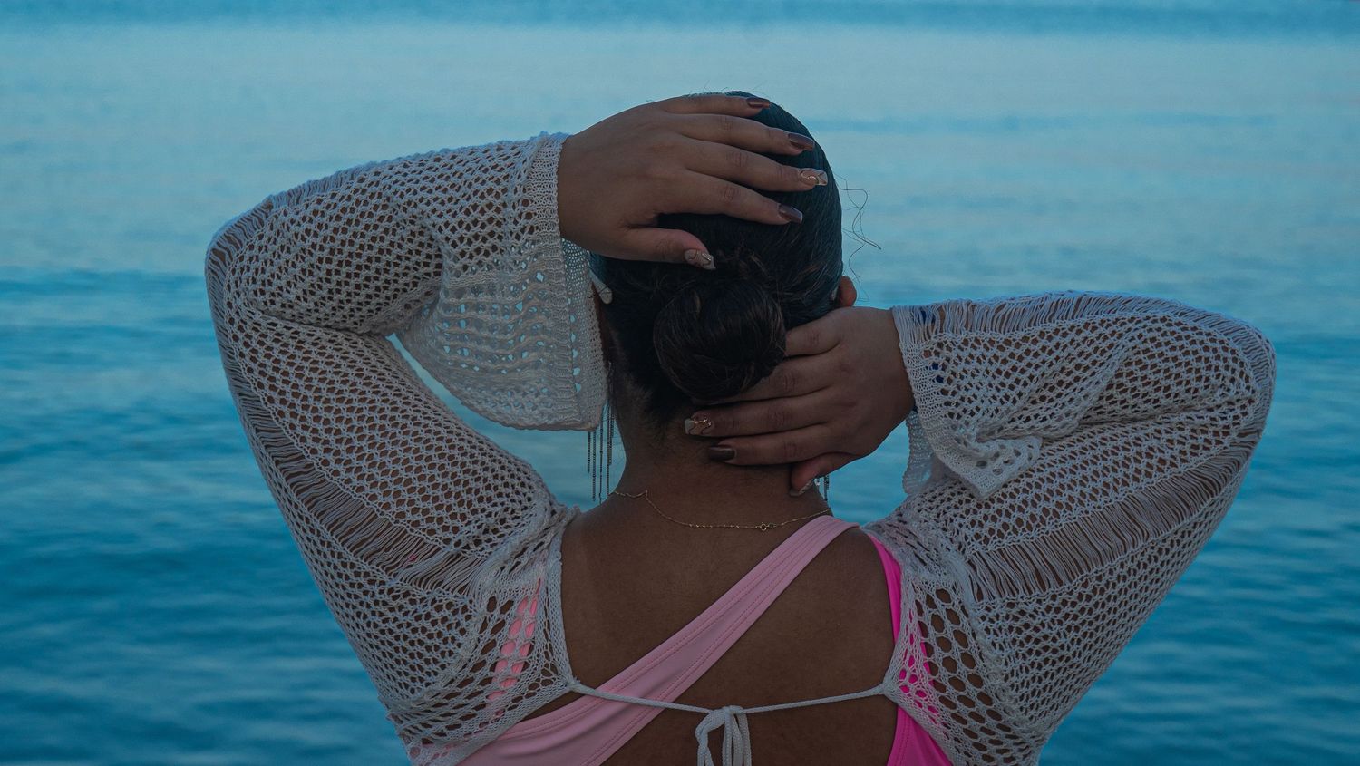 Person wearing a white mesh top and pink swimsuit gazes at the ocean on a bright blue summer day.