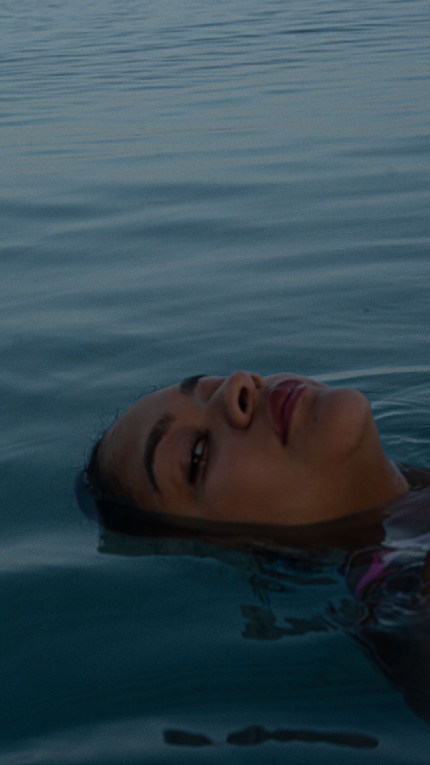 Figure floating peacefully in dark blue water at dusk, with rippling reflections on the calm surface.