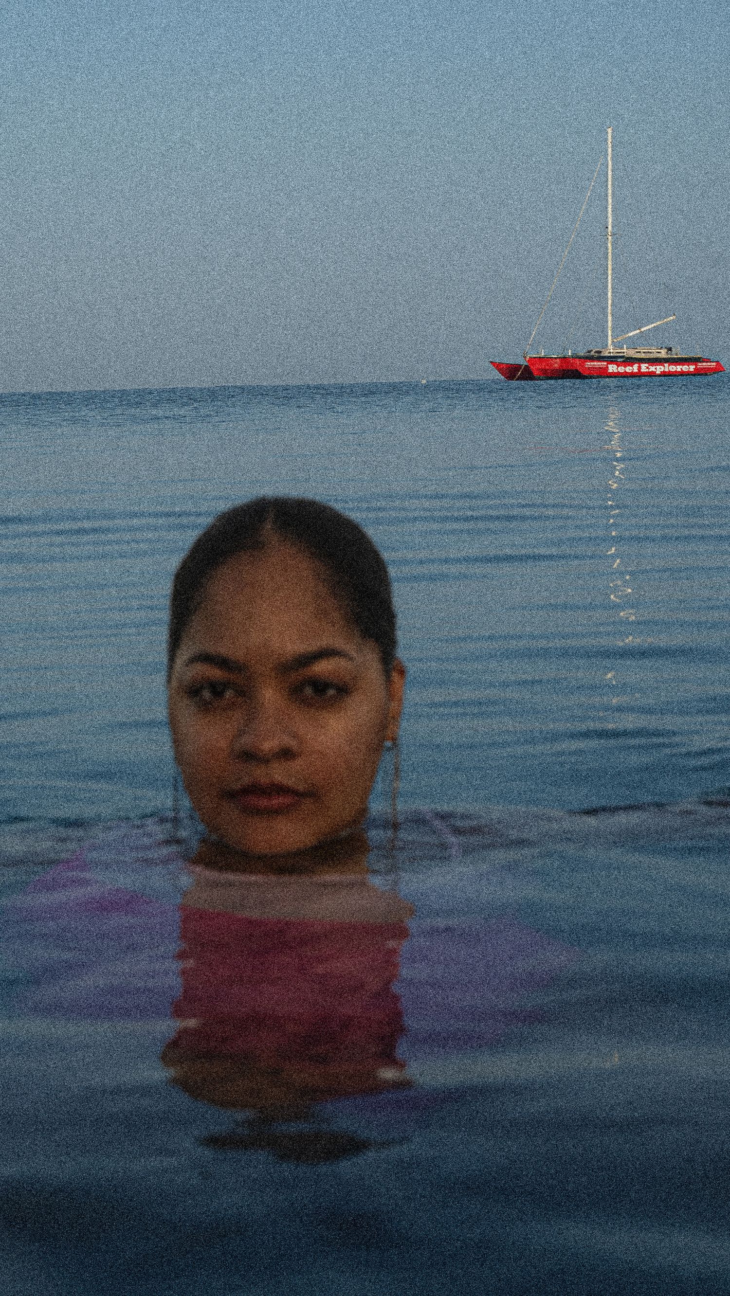 A swimmer floats in calm blue waters with a red sailboat visible in the distance on the horizon.