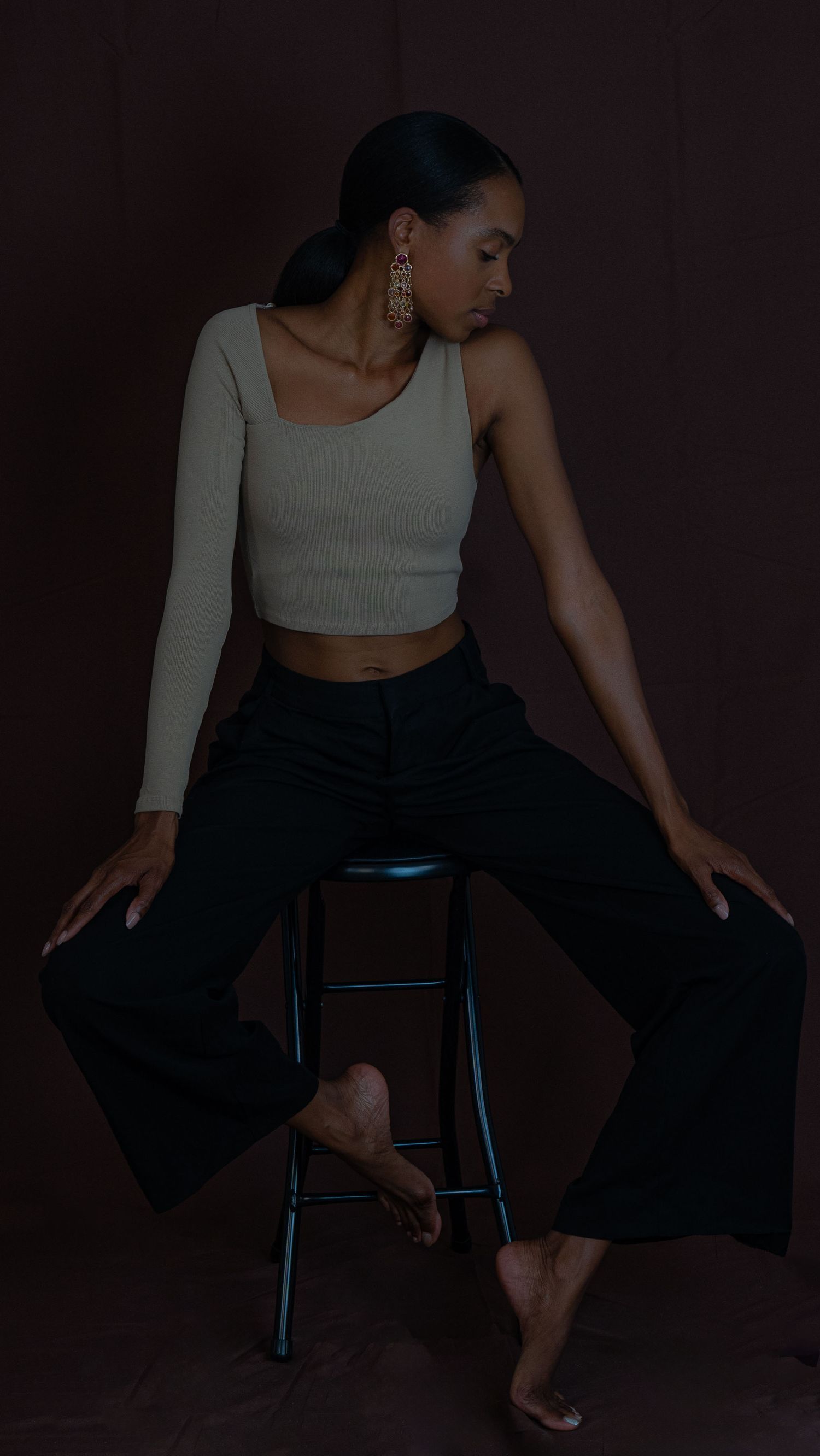 Artistic portrait of model in asymmetrical crop top and wide-leg pants posing elegantly on dark stool against brown backdrop.