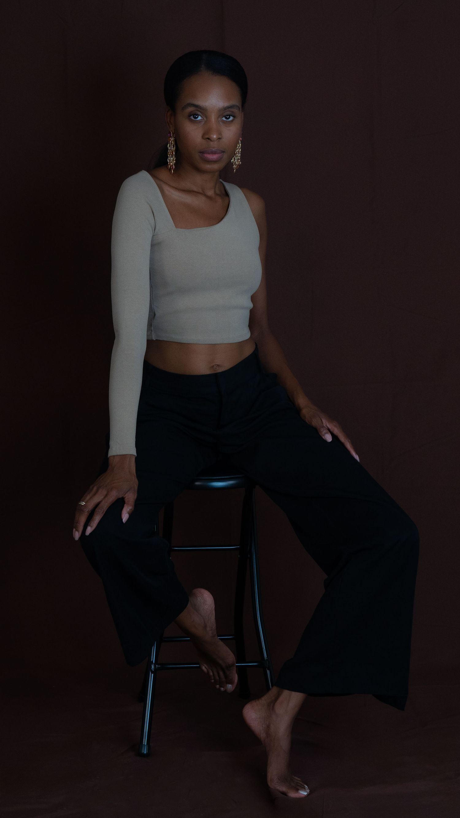 Elegant model in gray crop top and black pants poses on stool against dark brown backdrop for fashion photoshoot.