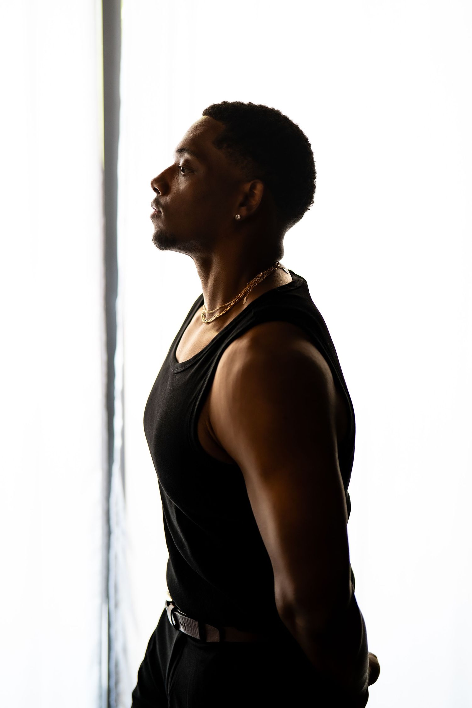 Young Black man in black tank top and gold chain necklace posed in dramatic backlit silhouette by window.
