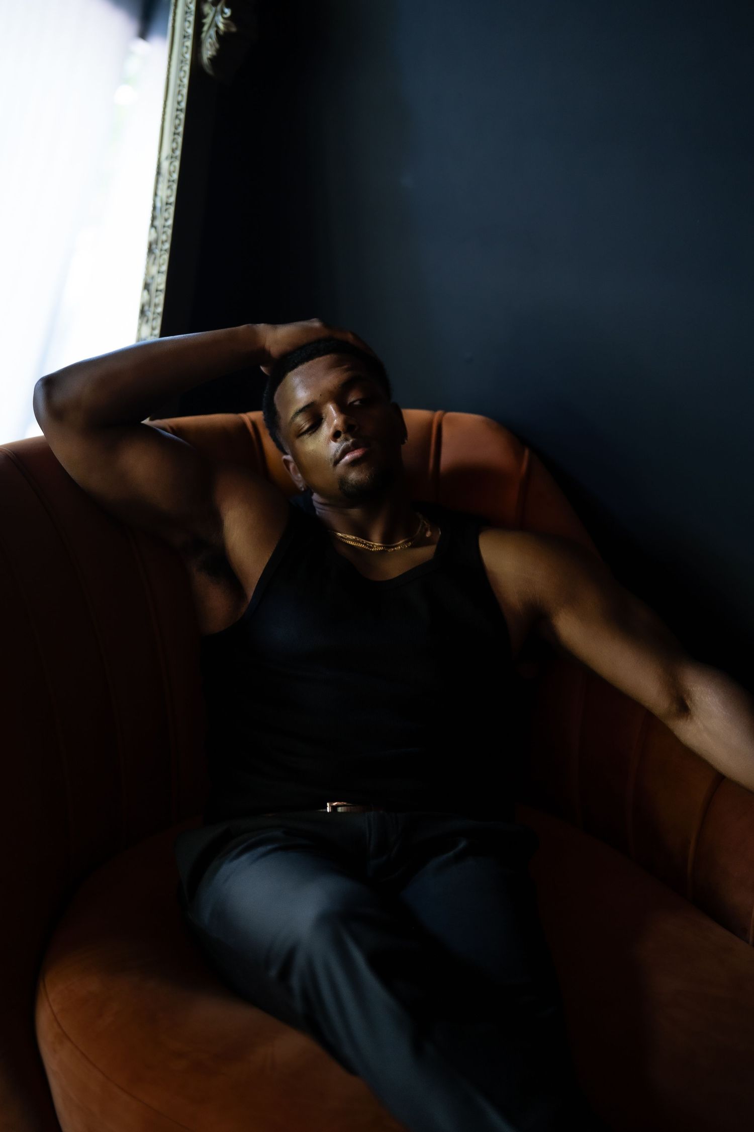 A muscular Black man in a black tank top and dark jeans relaxes on a brown leather sofa against a dark wall.