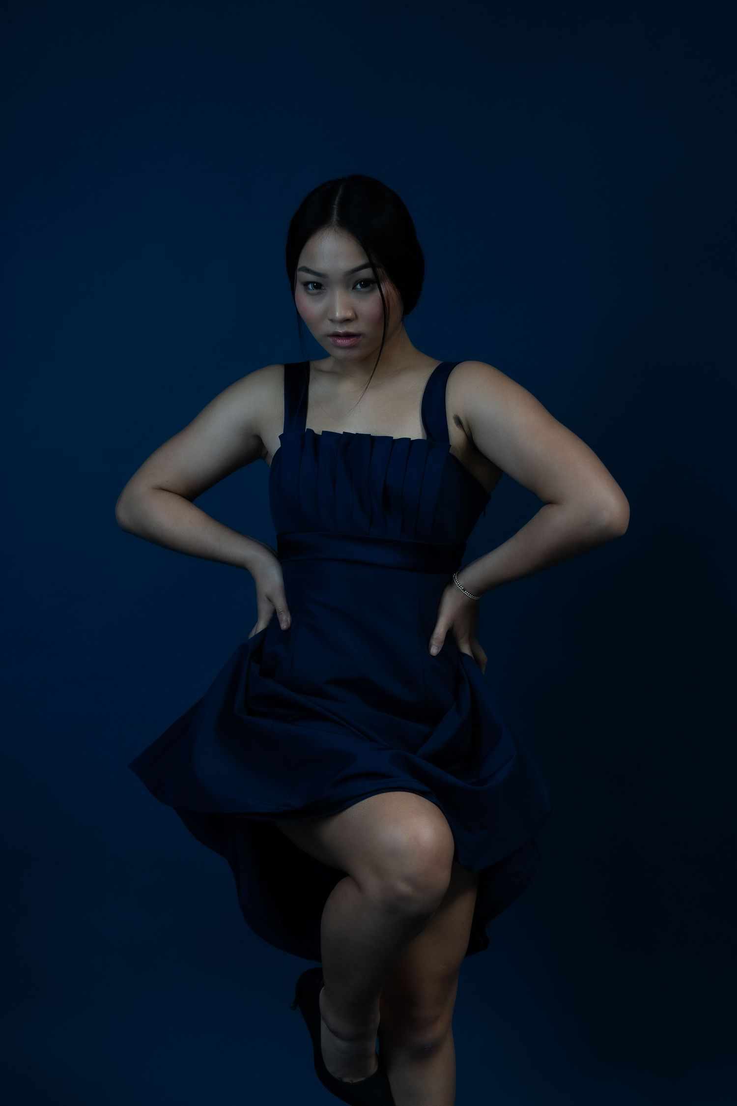 Model in navy dress strikes dynamic pose against deep blue backdrop in moody lighting.