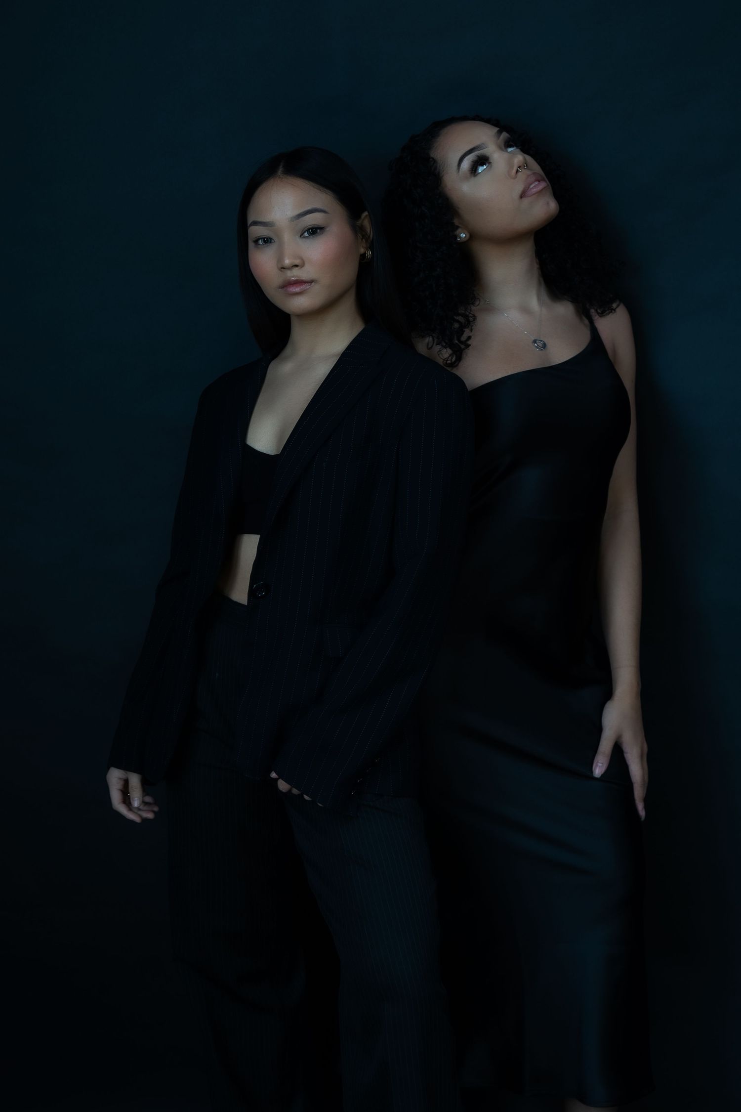Two models in dark outfits pose dramatically against black background.