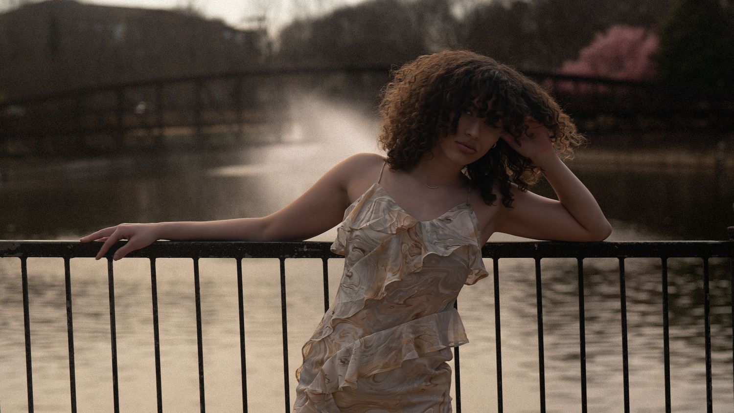 A person in a floral dress poses gracefully on a wooden deck by a serene lake at sunset.