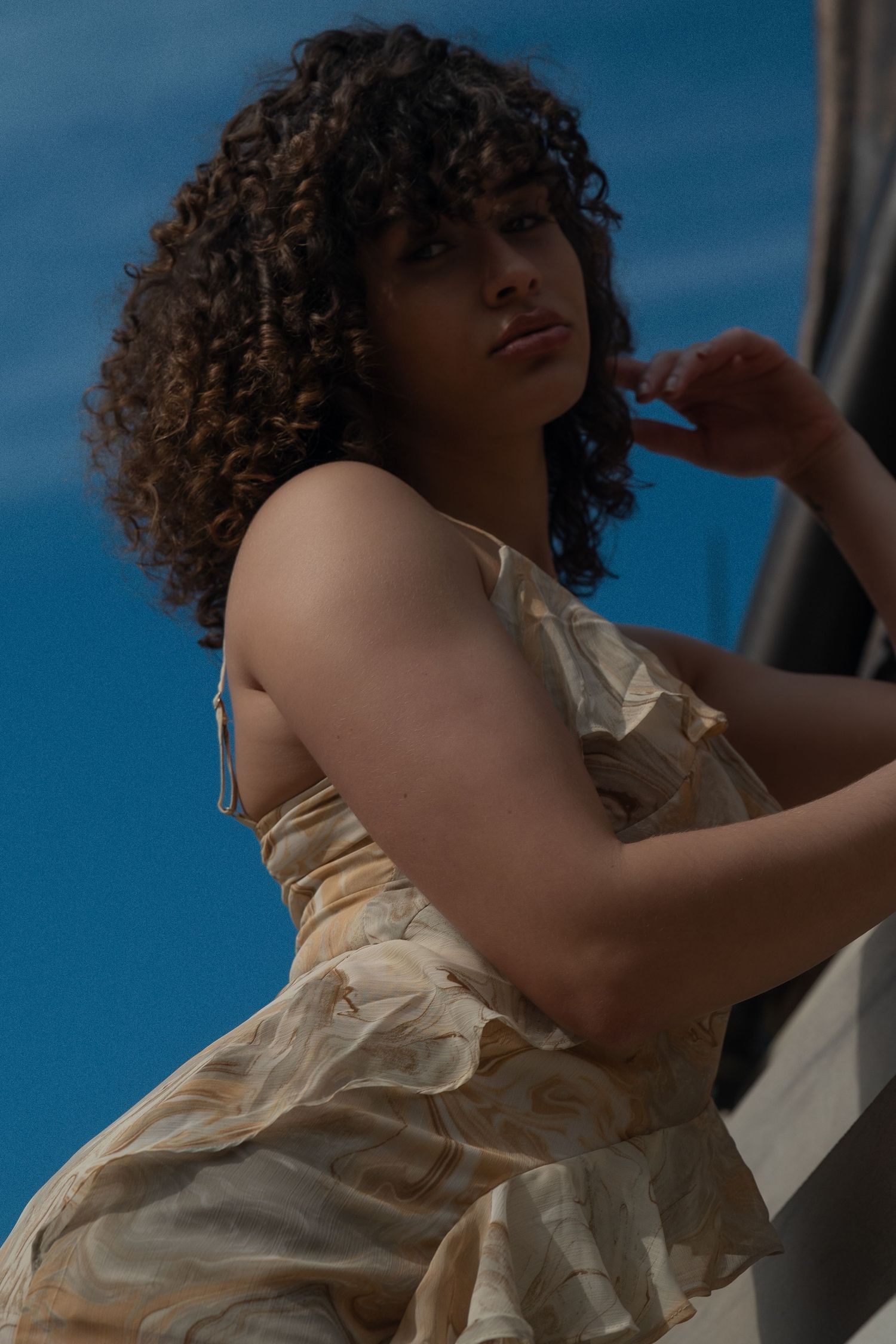 A person with curly hair in a cream colored top poses against a blue background in an artistic photo series.