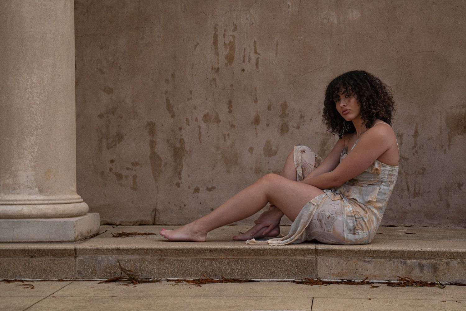 Woman with curly hair in slip dress sits barefoot against weathered stone wall and column.