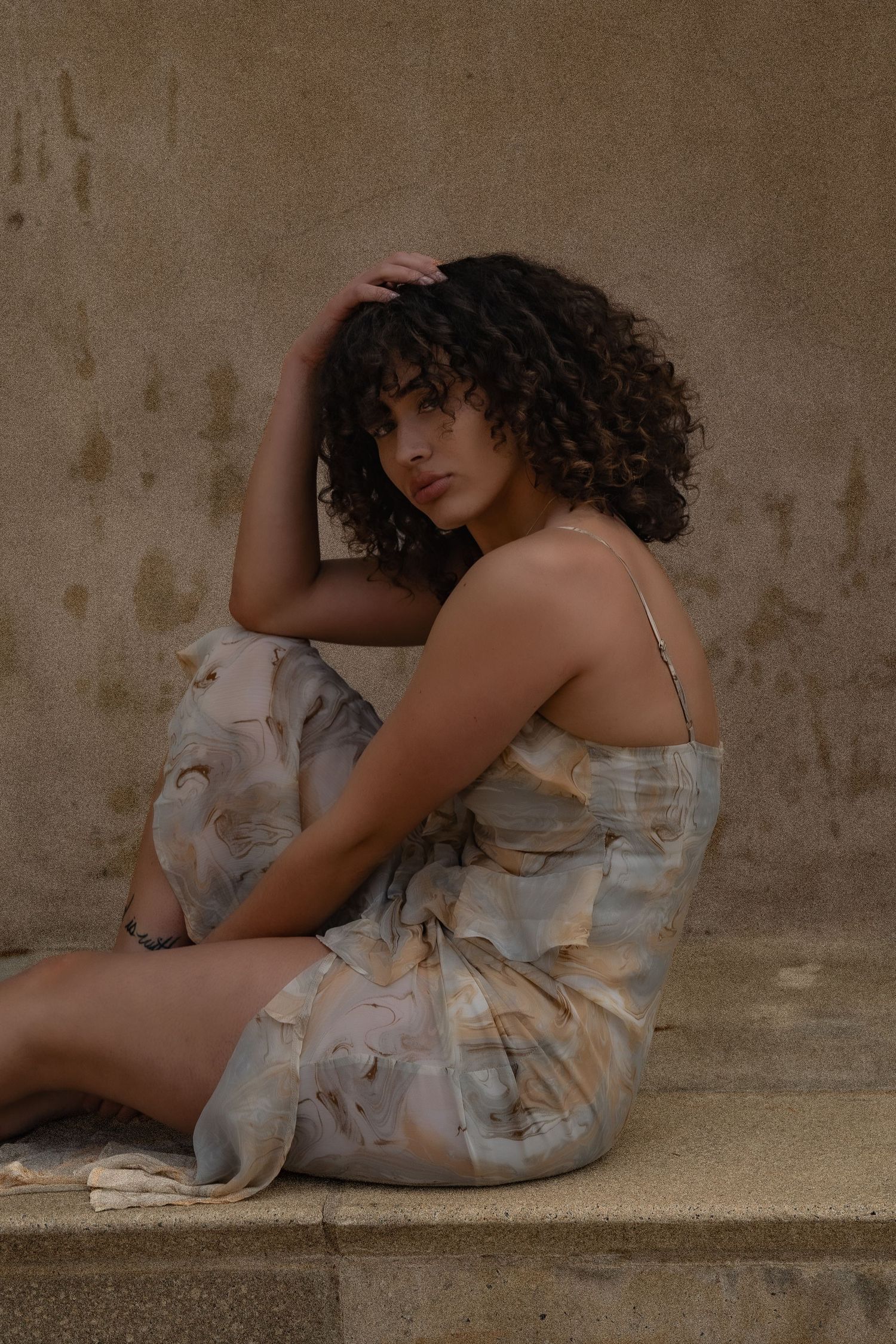 Portrait of person with curly hair wearing a floral sundress sitting against textured wall in moody lighting.