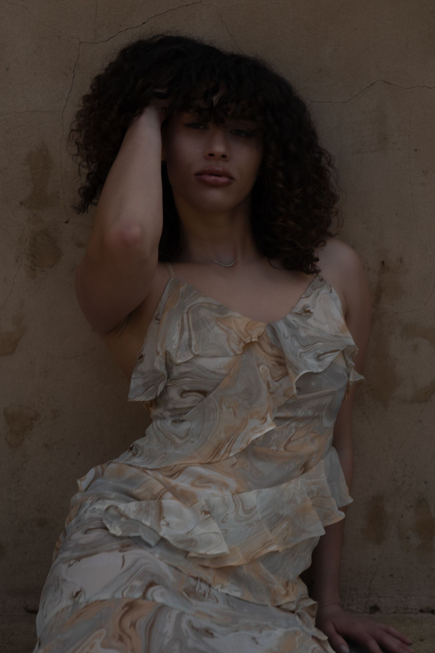 Figure in white ruffled dress poses artistically against dark wall with dramatic shadows.