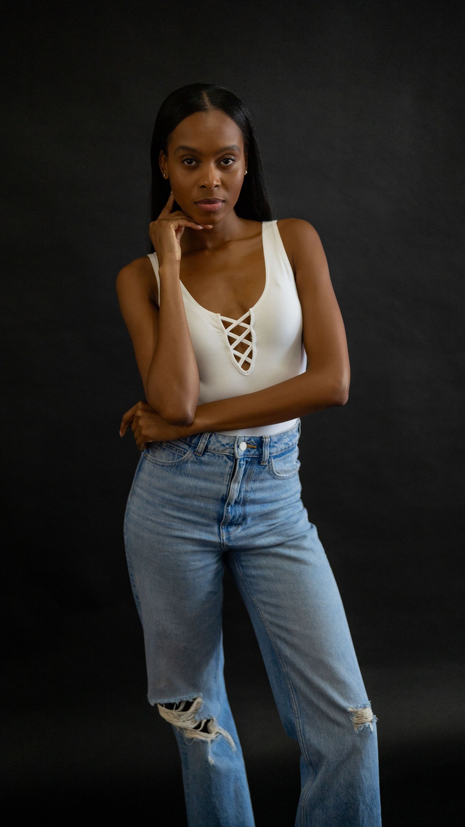 Person posing against dark background wearing white lace-up bodysuit and distressed light blue jeans.