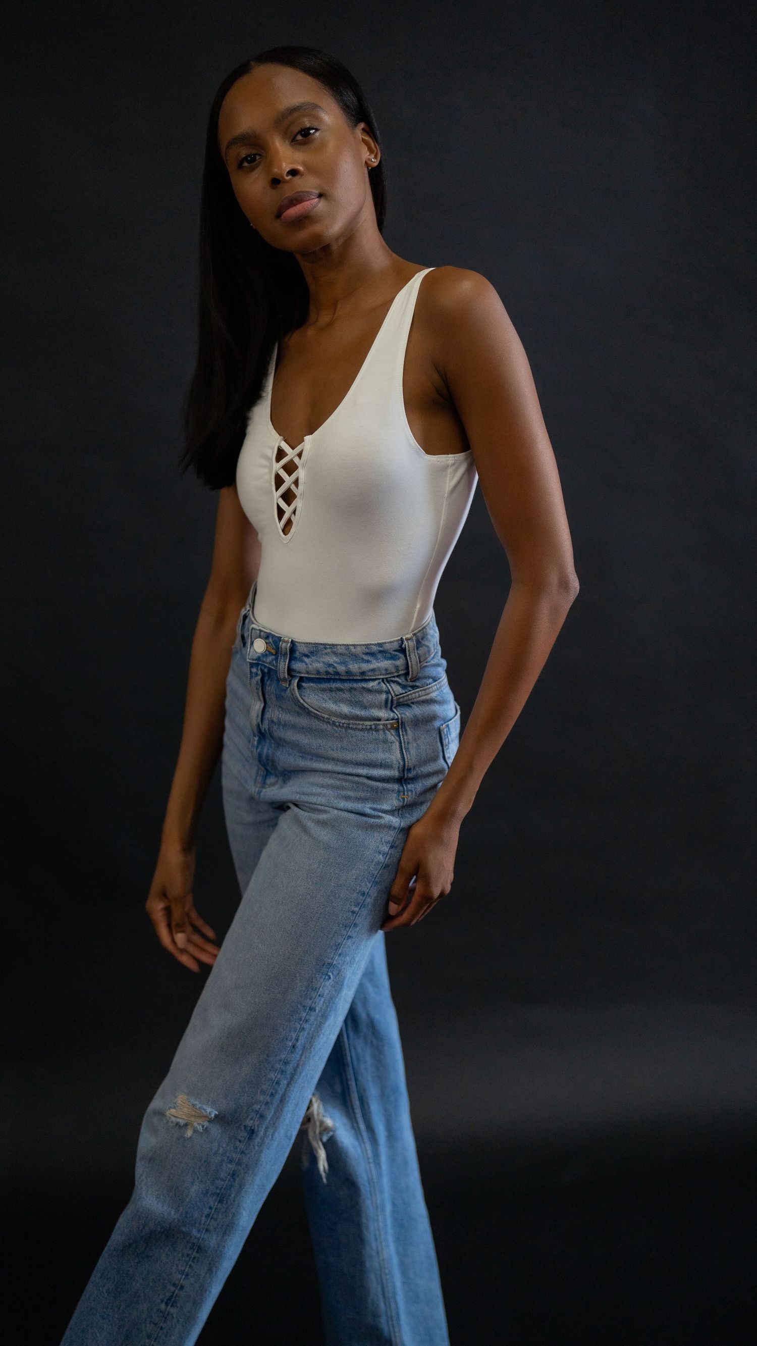 Fashion model wearing a white lace-up bodysuit and distressed light blue jeans poses against a dark background.