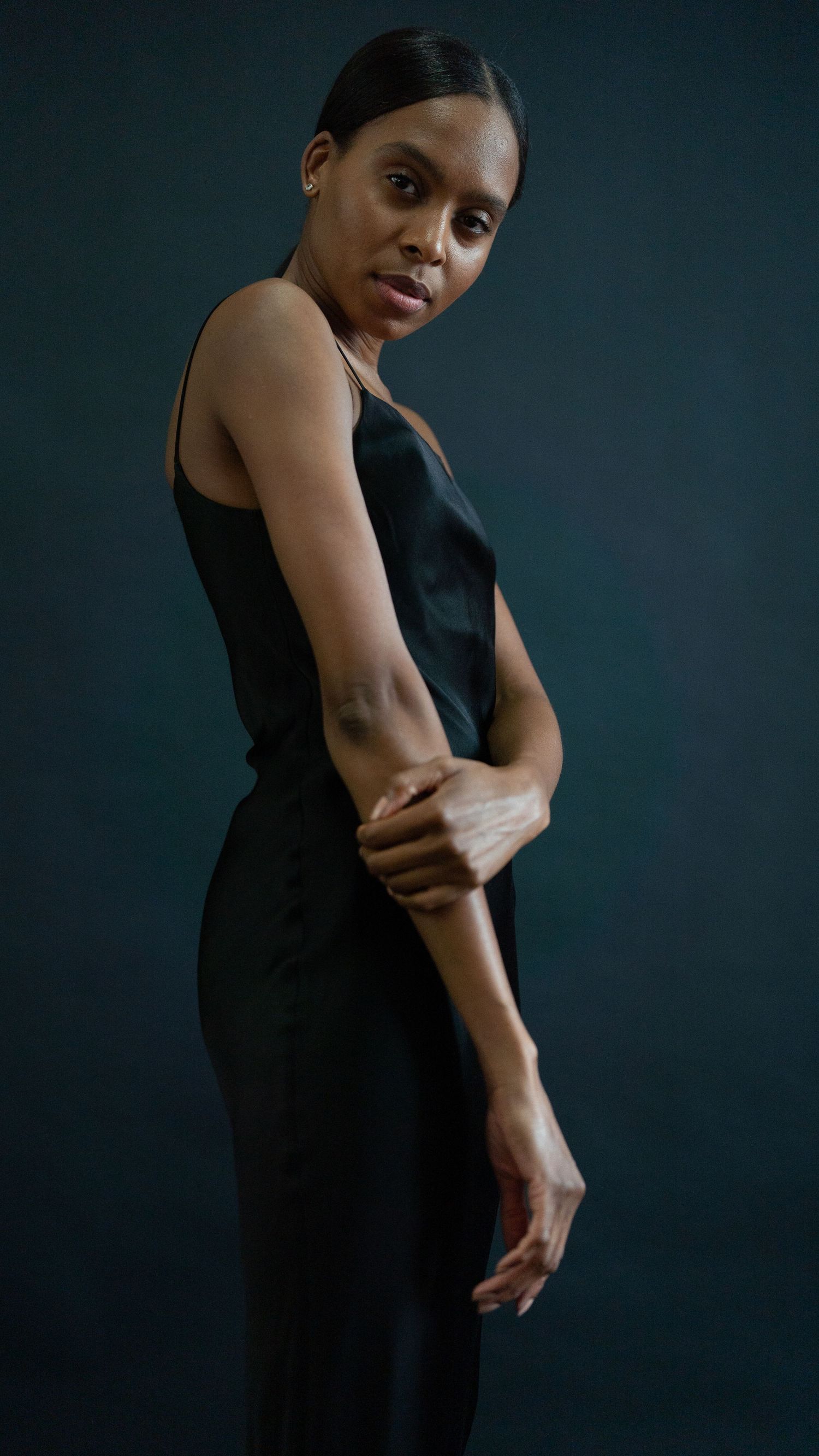 A woman in an elegant black sleeveless dress poses gracefully against a dark background for an artistic portrait.