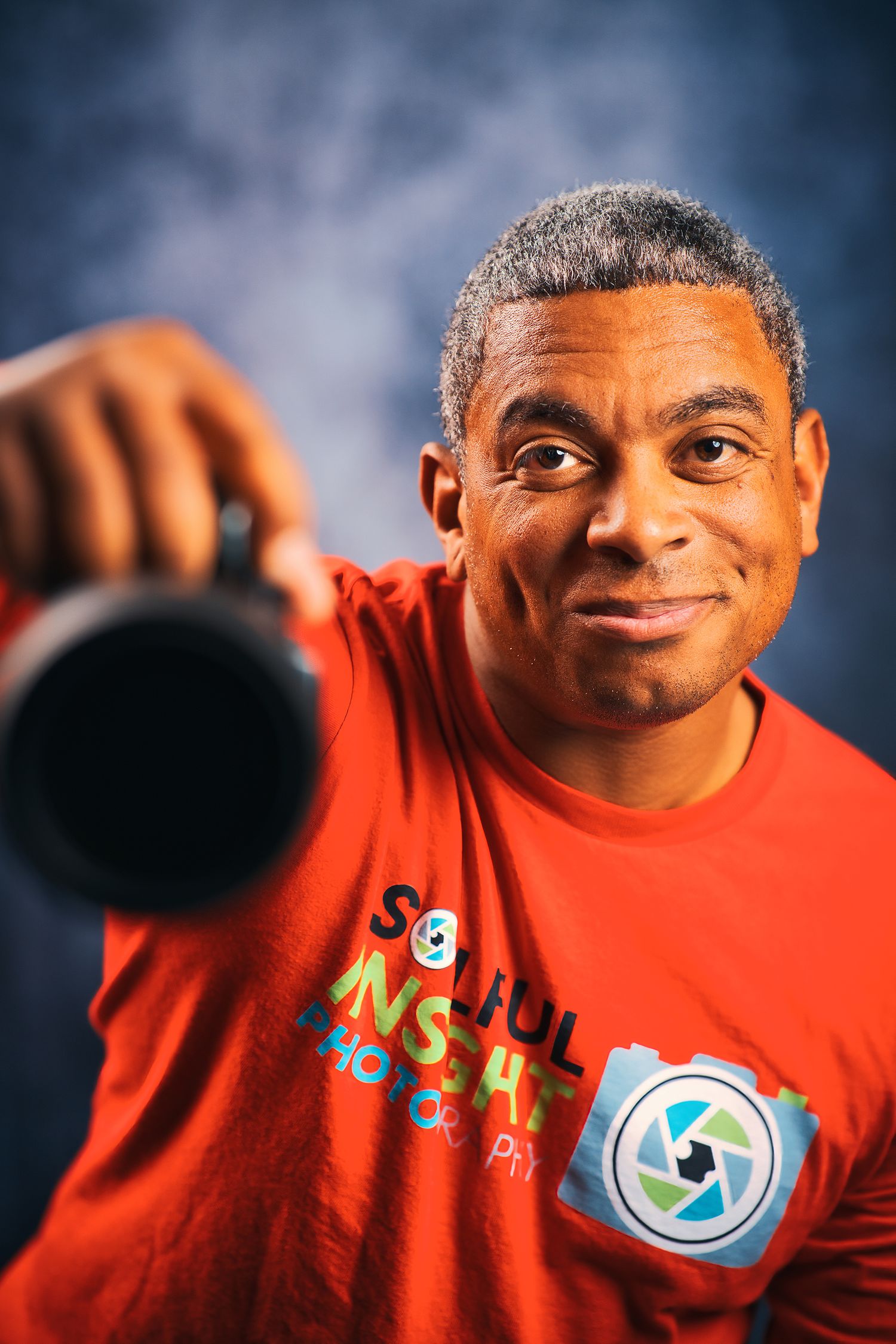 Portrait of photographer in red shirt holding camera lens against blue background.