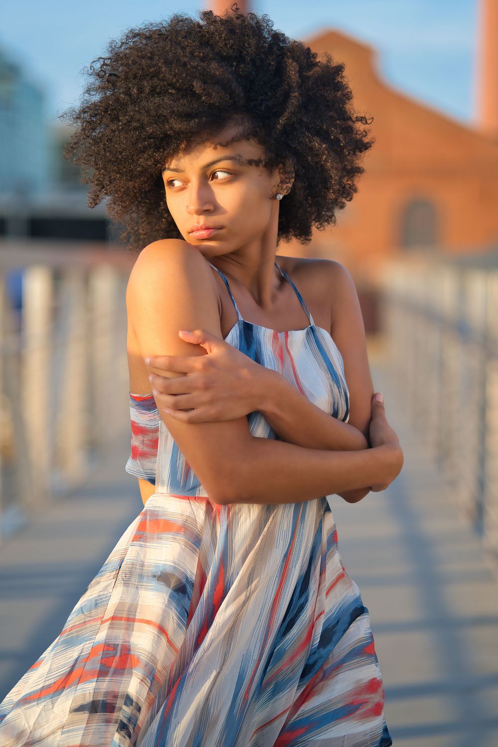 Young Black woman with afro hair in striped sundress hugging herself on a bridge at golden hour.