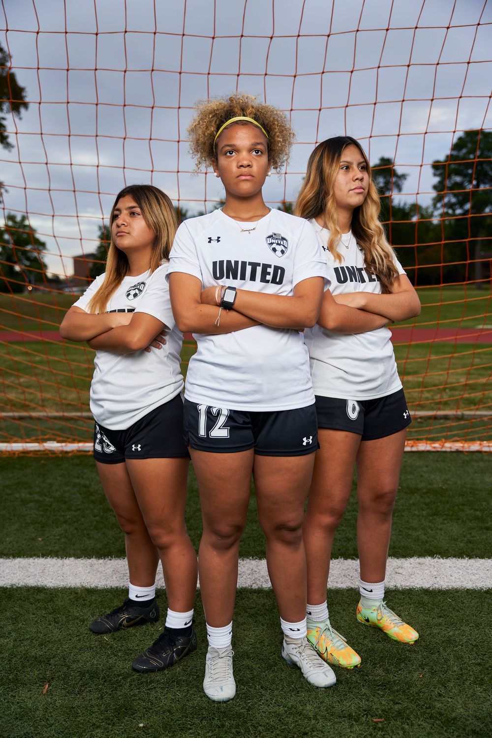 Three female soccer players in white United jerseys pose with arms crossed in front of a goal net on a field.