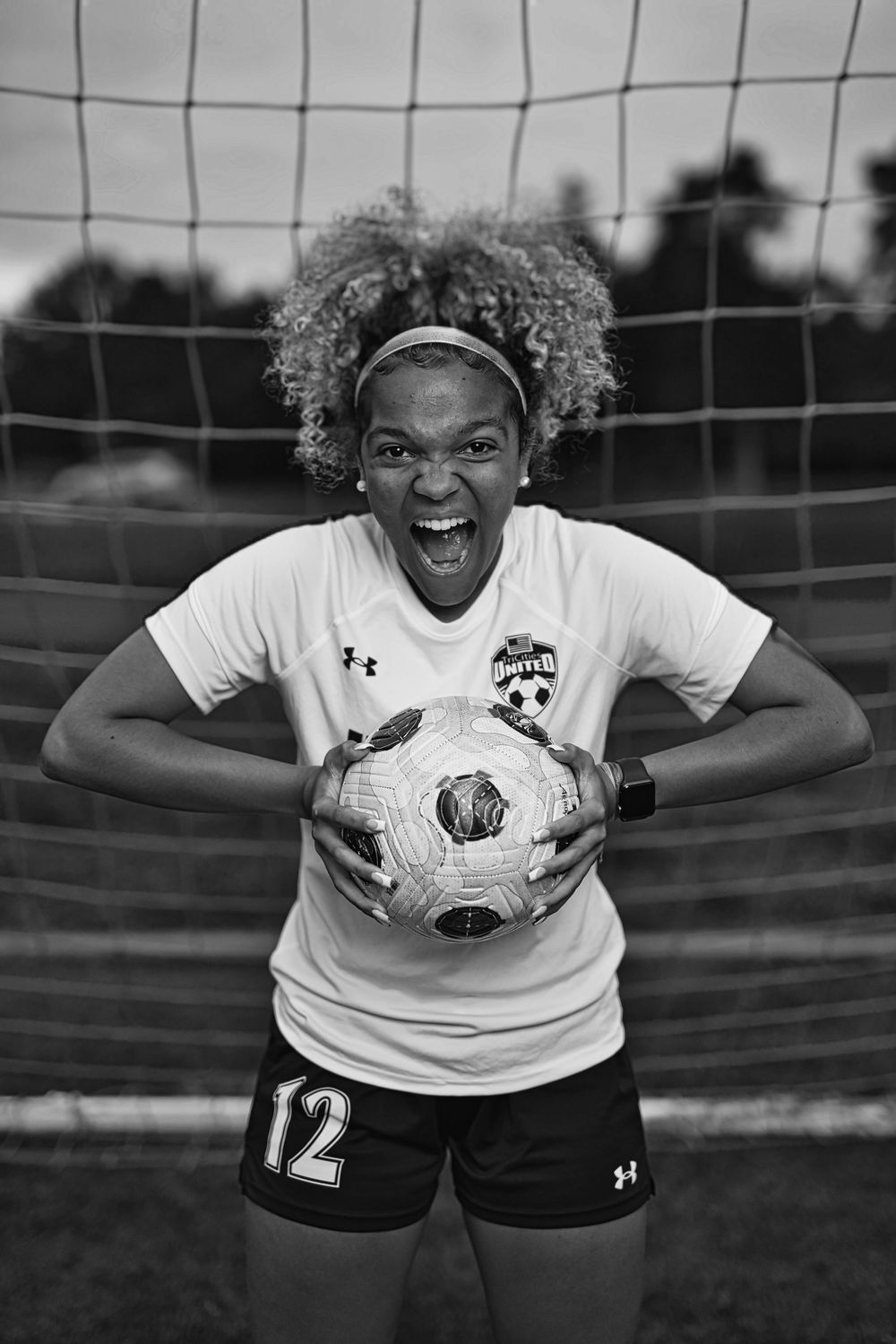 Young female soccer player in jersey number 12 holds ball, roaring confidently in front of a goal net.