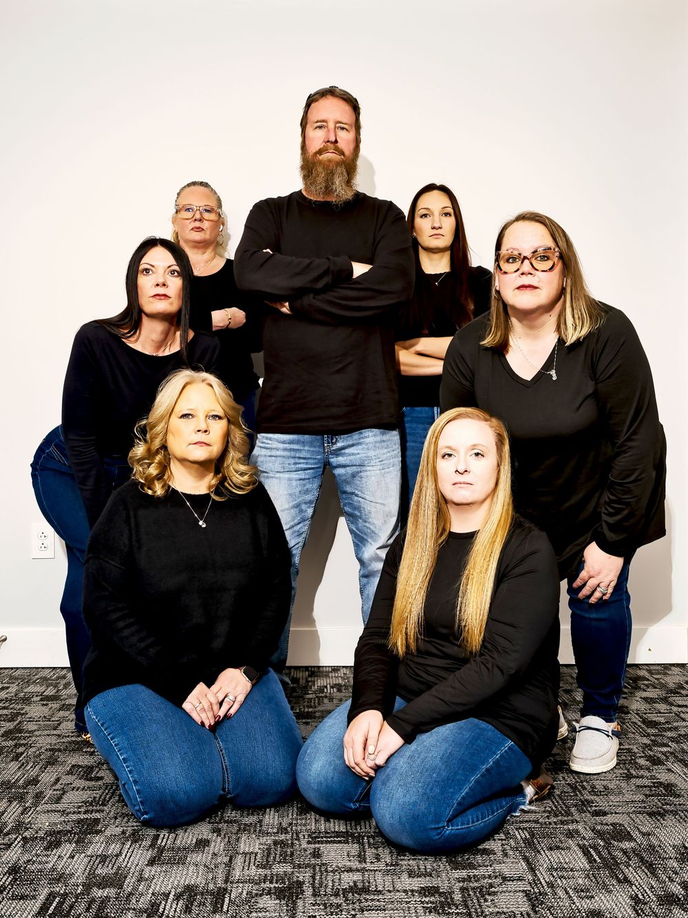 Group of seven adults in black tops and jeans posing together, some seated on floor, against white wall.