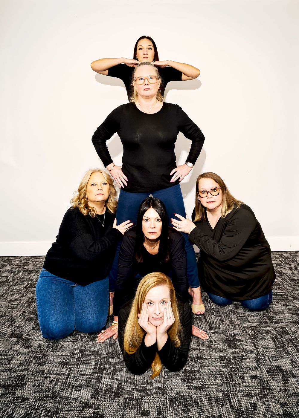 Five women in black tops pose in a pyramid formation against a white wall on a carpeted floor.