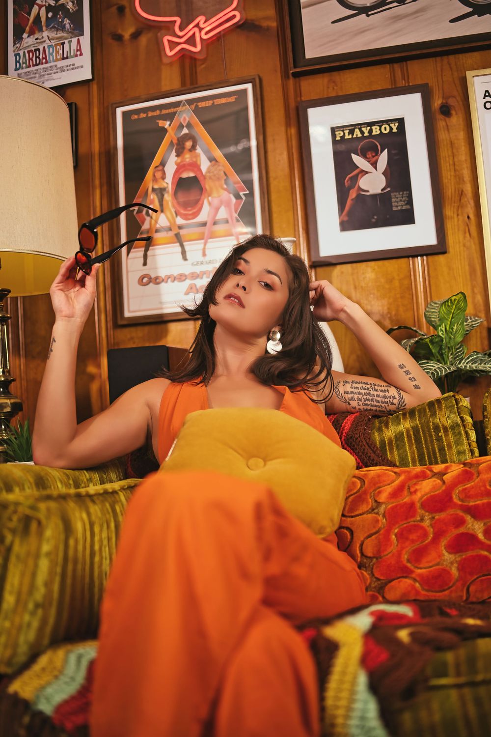 Woman in orange outfit sitting on retro couch surrounded by vintage movie posters in a 70s-inspired room.