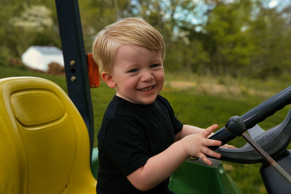 Happy toddler boy smiling while playing on a green and yellow toy tractor outdoors.