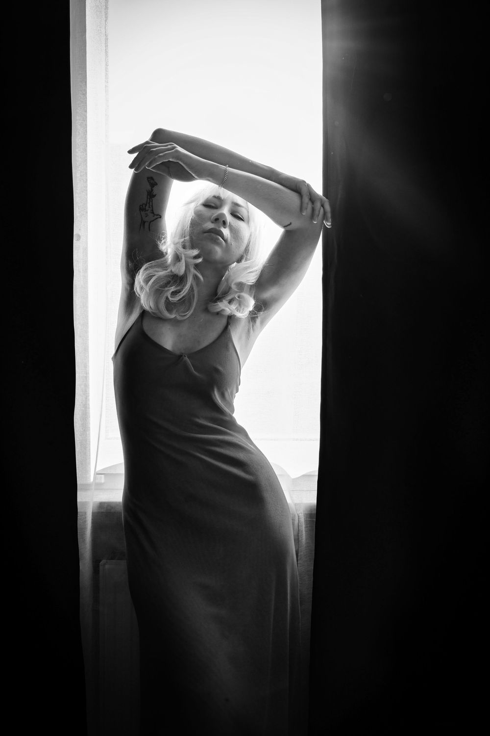 Black and white photo of a blonde woman in a slip dress posing gracefully by a bright window with arms raised.